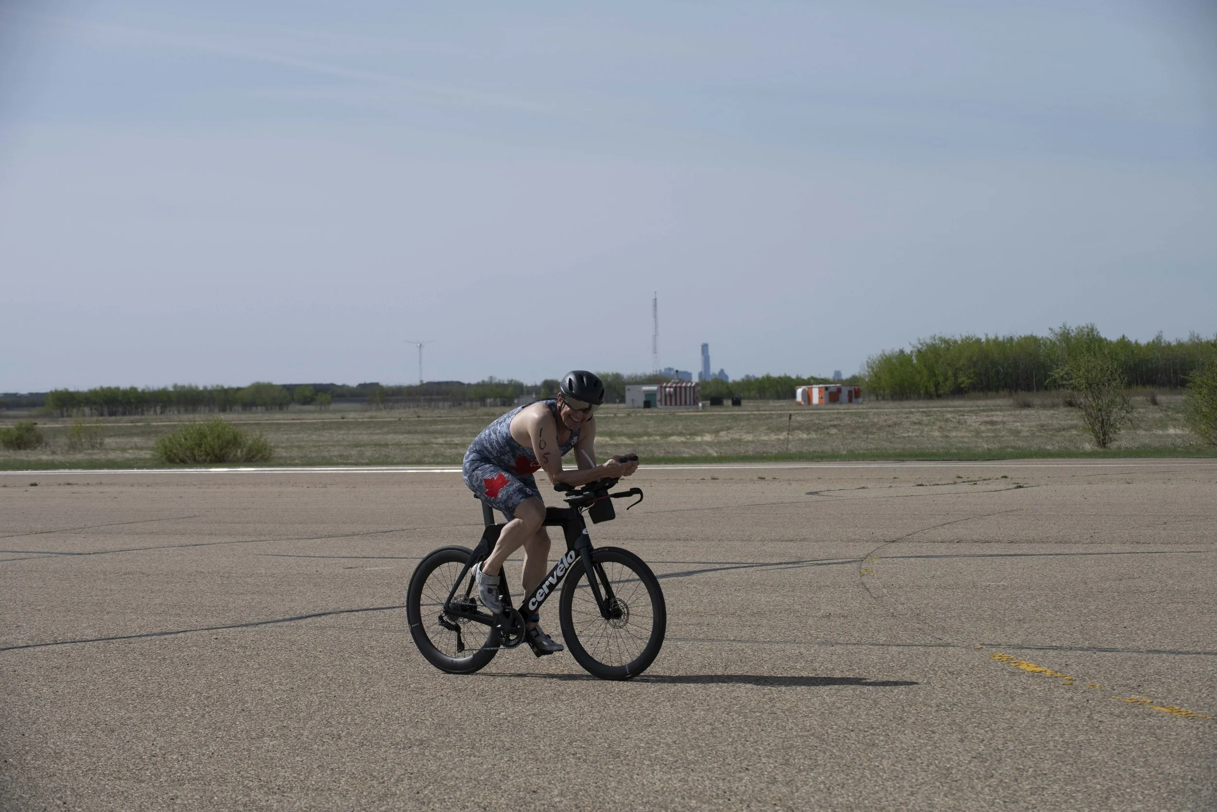 A person riding a black bicycle on an open paved area, wearing a helmet and athletic clothing with a red cross on the shorts, smiling and looking down at a device in their hand.