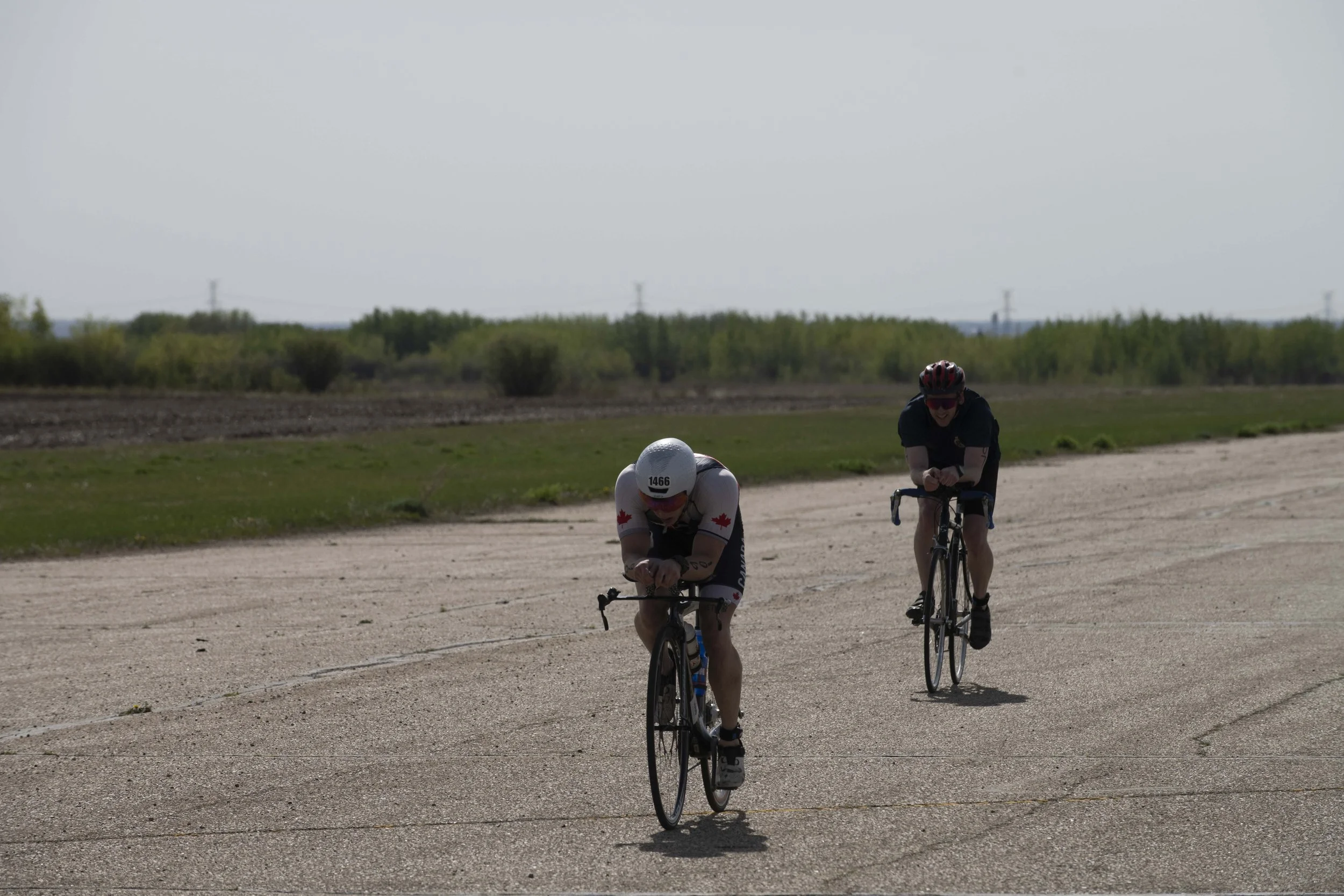 Two cyclists riding on a flat, open road in a rural area with green fields and trees in the background on a cloudy day.