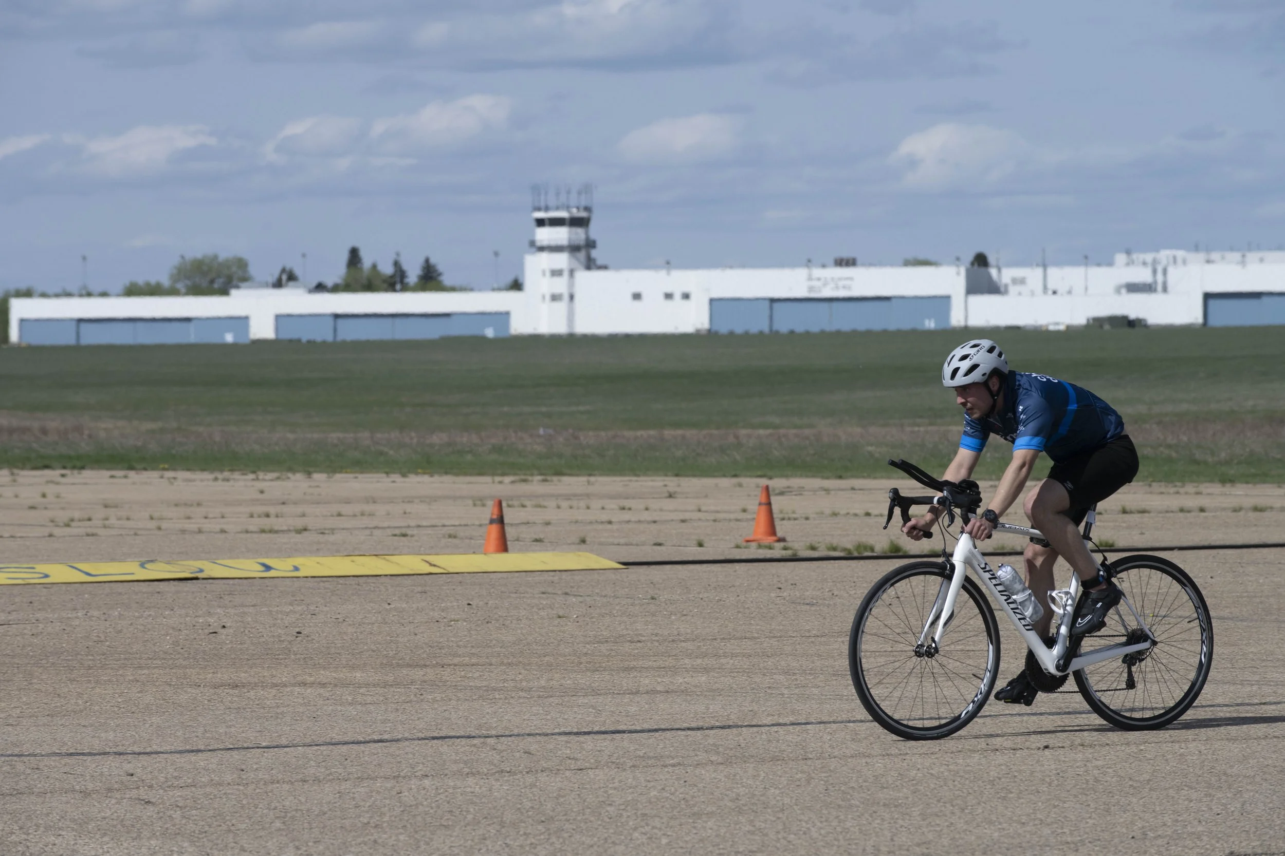 A person wearing a blue shirt, black shorts, and a white helmet riding a white bicycle on a tarmac road, with orange traffic cones and a yellow line marked 'SLOW' in the foreground. An airport terminal building with hangars and control tower is visib