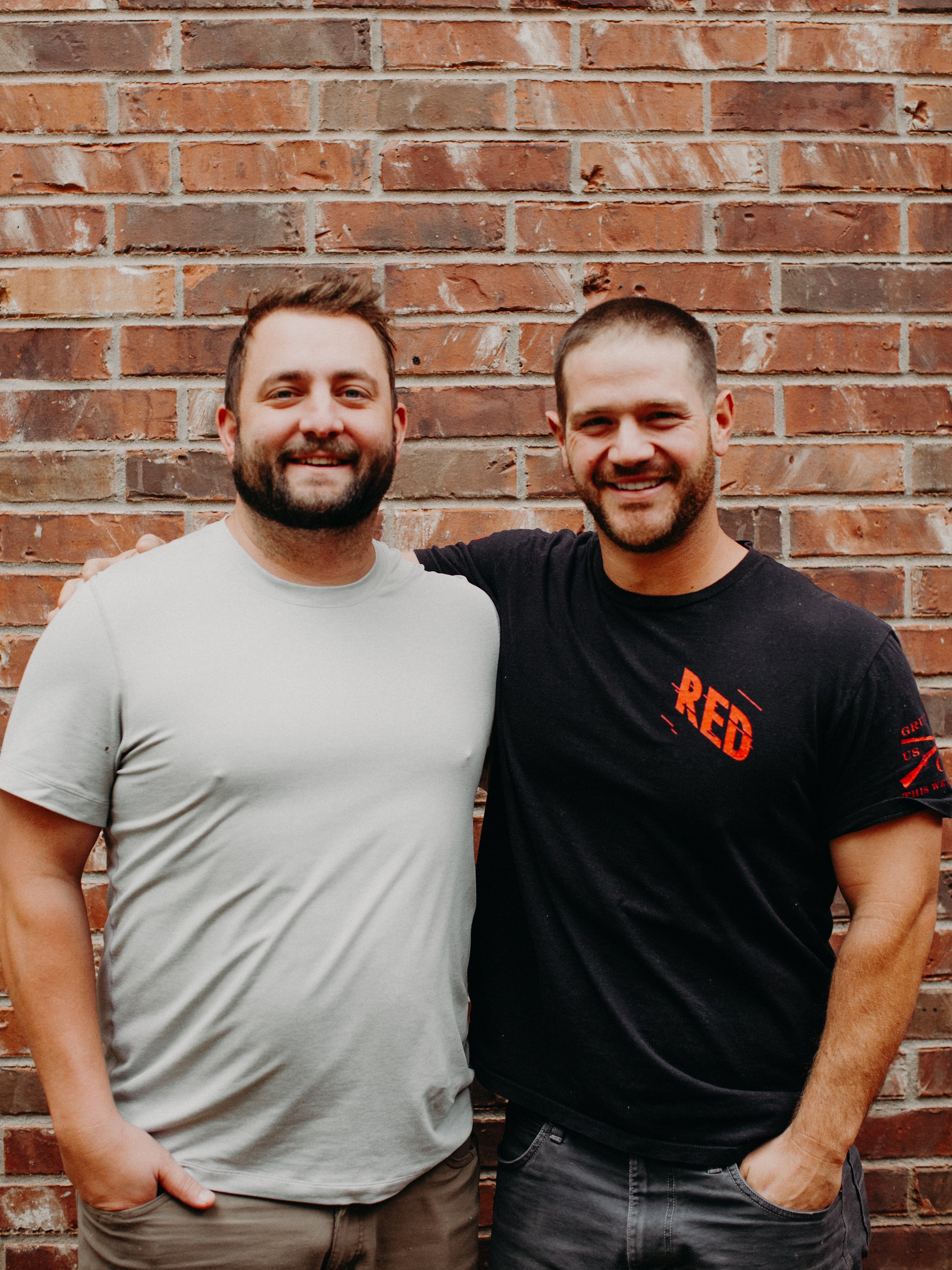 Two smiling men standing close together with their arms around each other's shoulders against a brick wall.