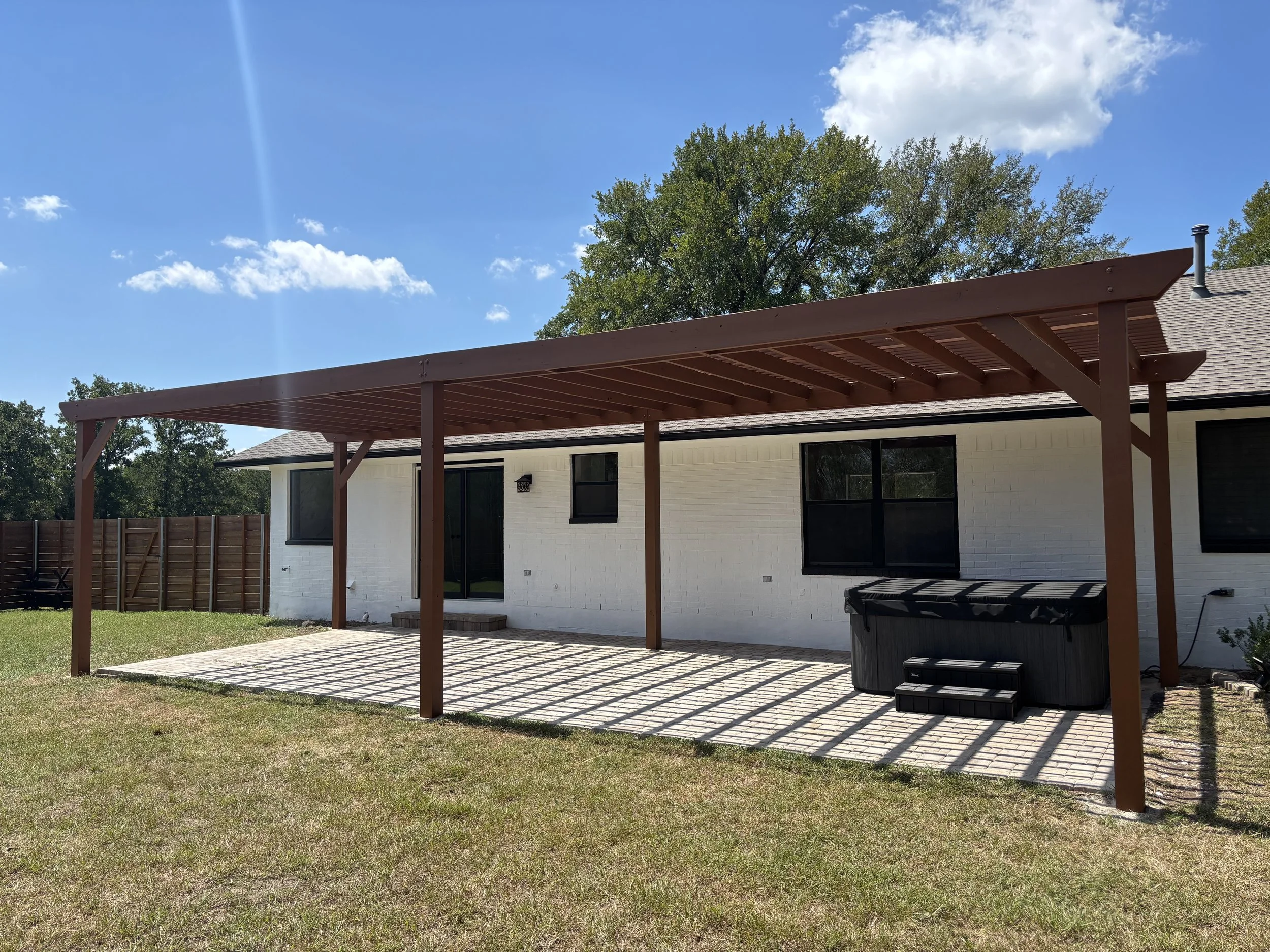 Backyard with wooden pergola, brick patio, hot tub, and white house with black-trimmed windows and sliding door under bright sunny sky.
