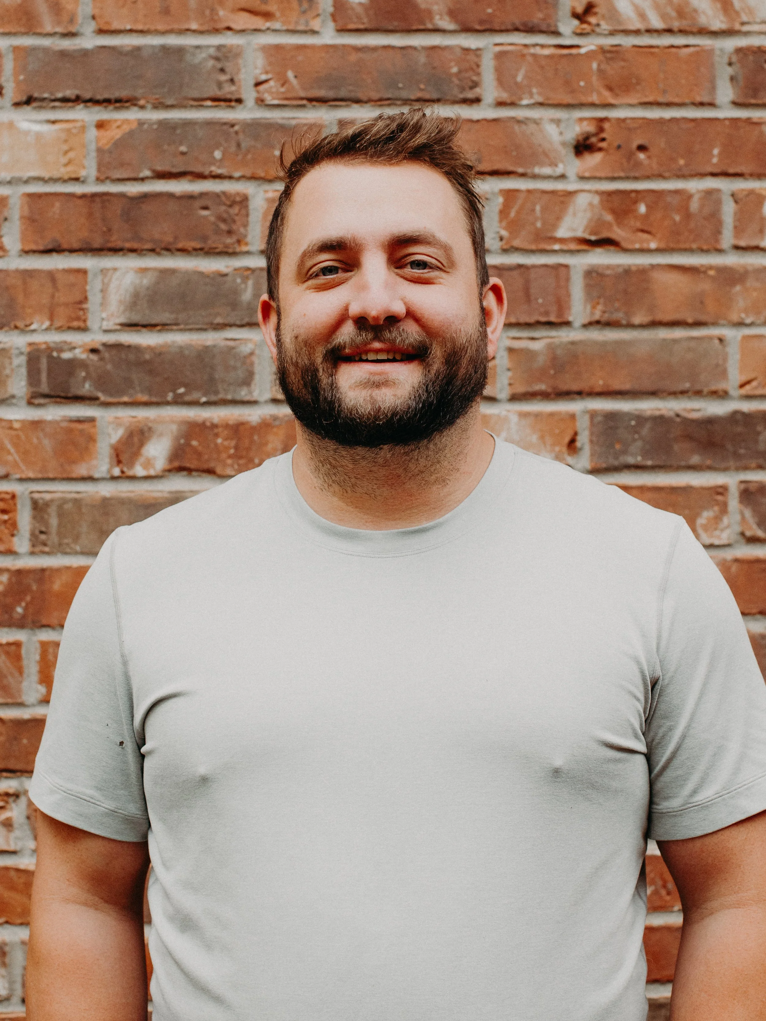 A smiling man with a beard and short brown hair standing in front of a brick wall, wearing a light gray t-shirt.