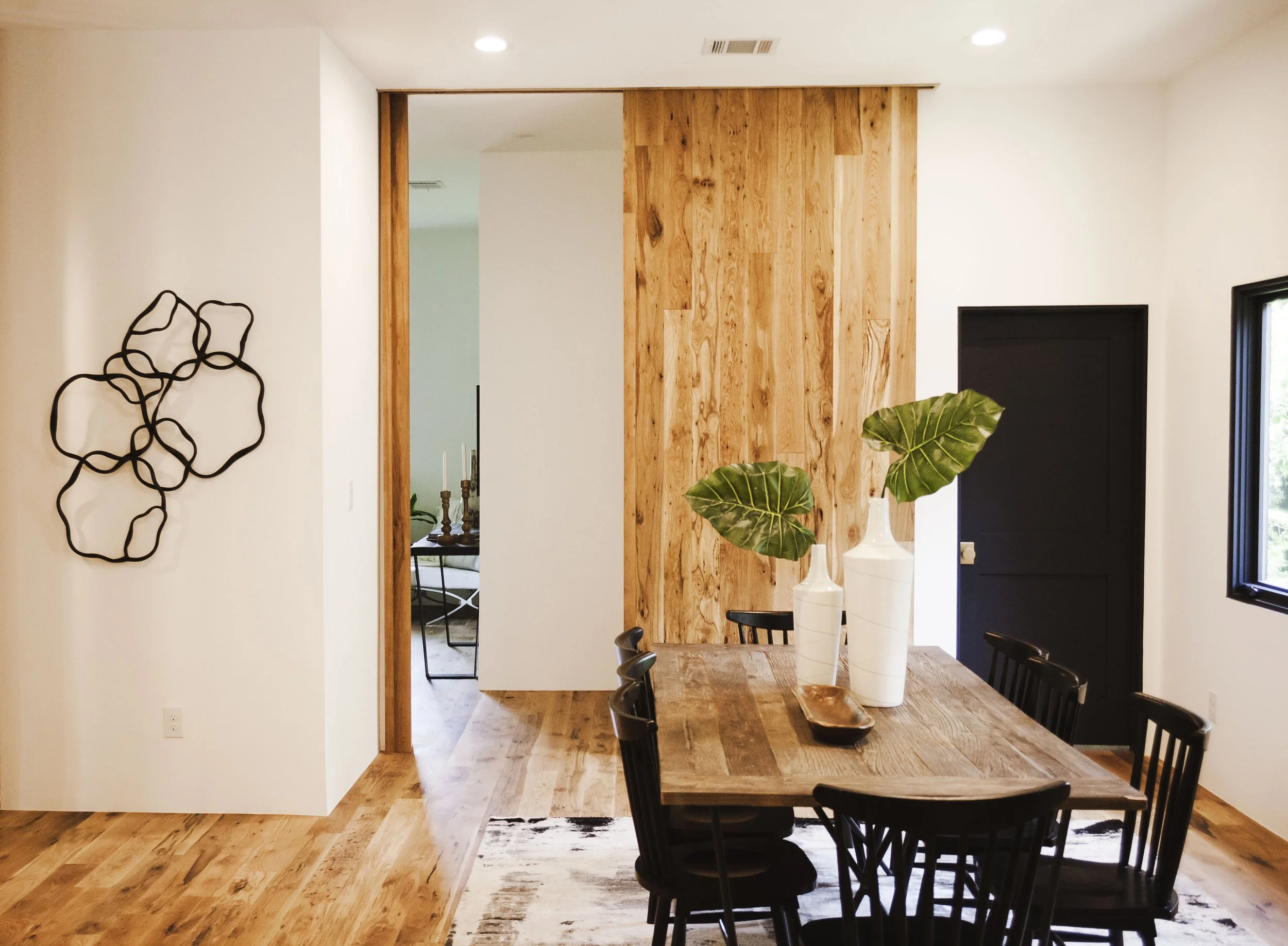 Modern dining room with a wooden table and six black chairs. Decor includes two large vases with leaves and a wall sculpture, with natural light coming through a window with black framing.