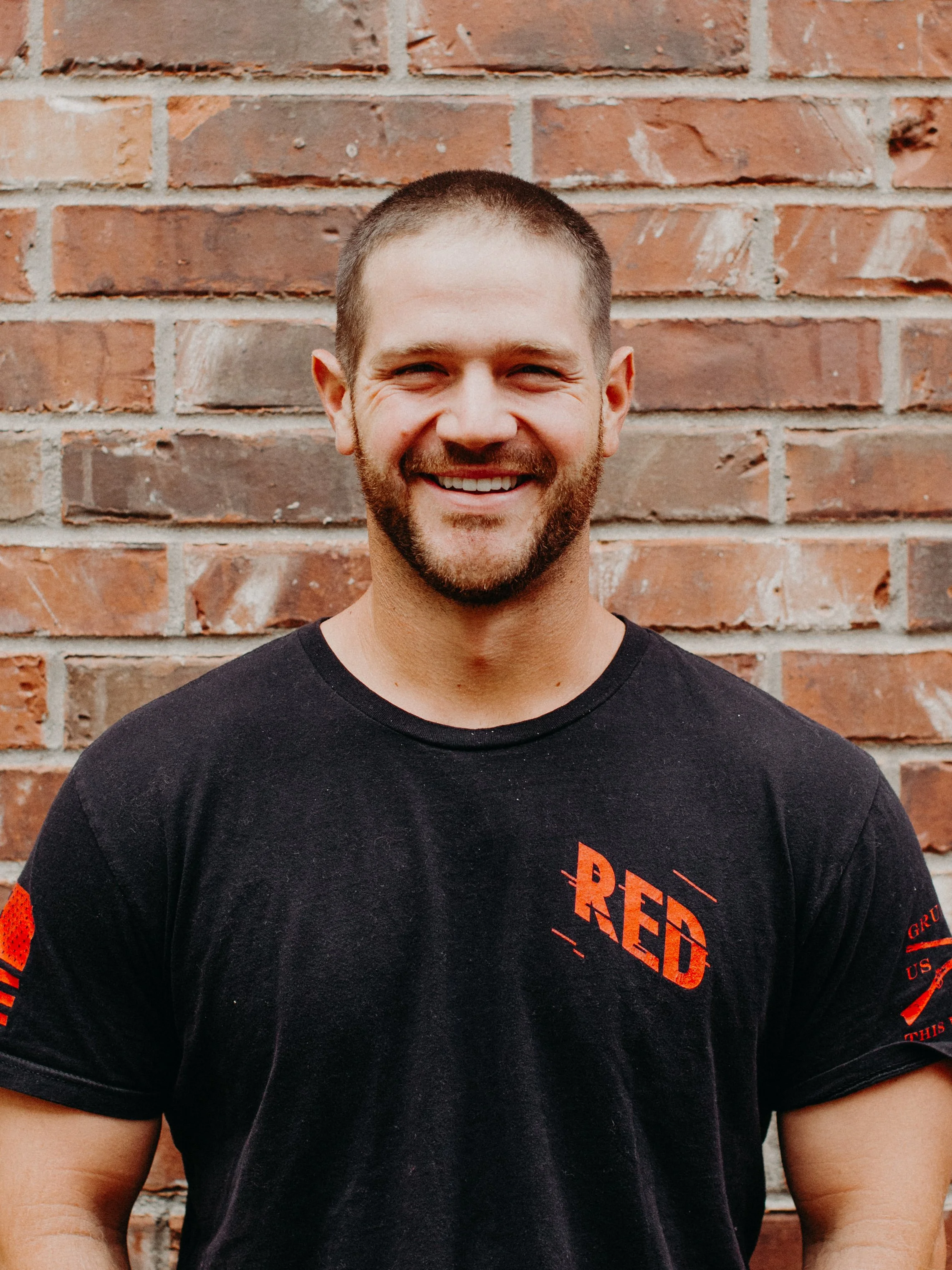 A smiling man with a beard and short hair, wearing a black t-shirt with the word 'RED' on it, standing in front of a brick wall.