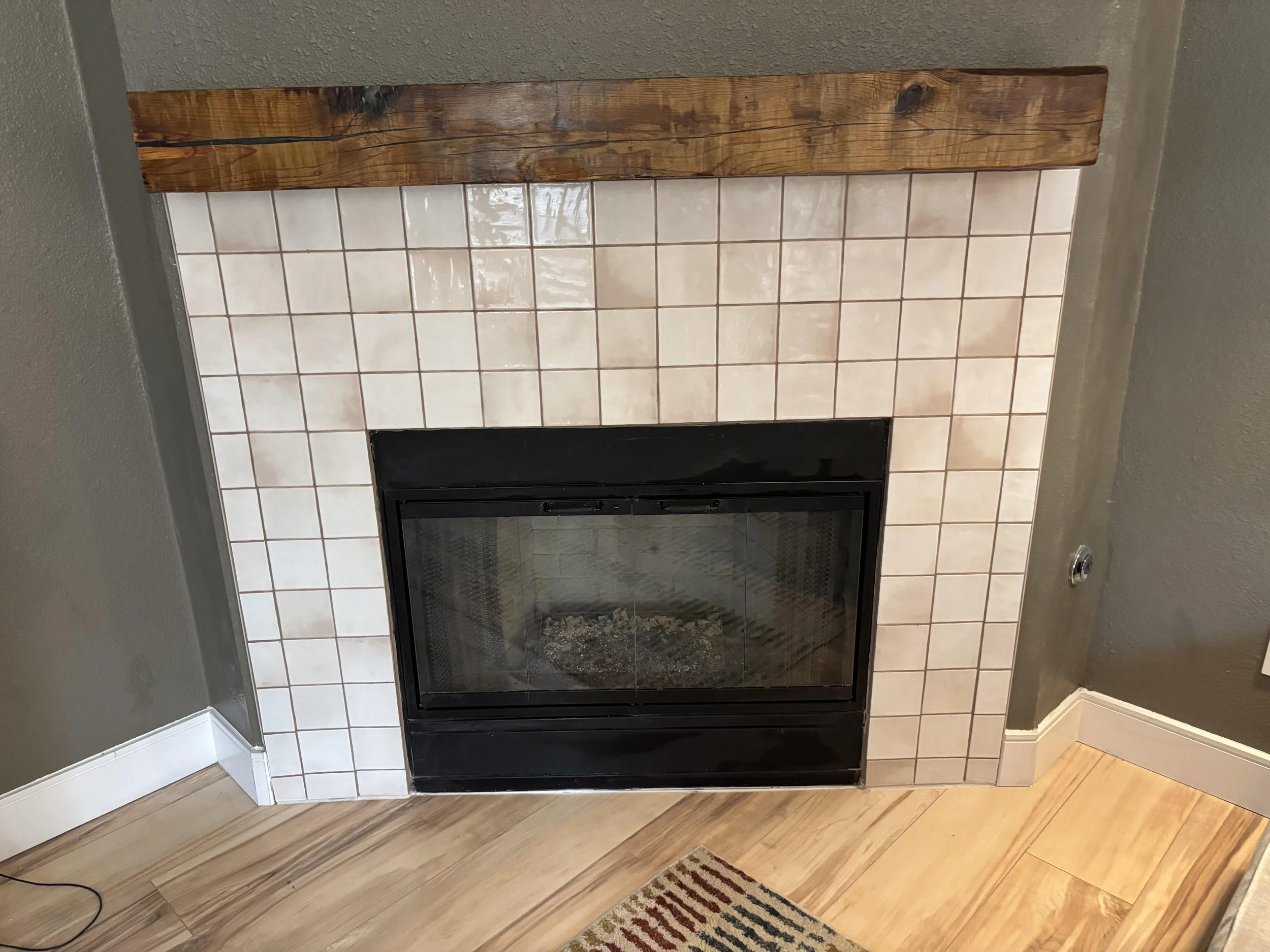 A fireplace with a black frame, surrounded by beige tiles, topped with a wooden mantel, in a room with green walls and hardwood flooring.