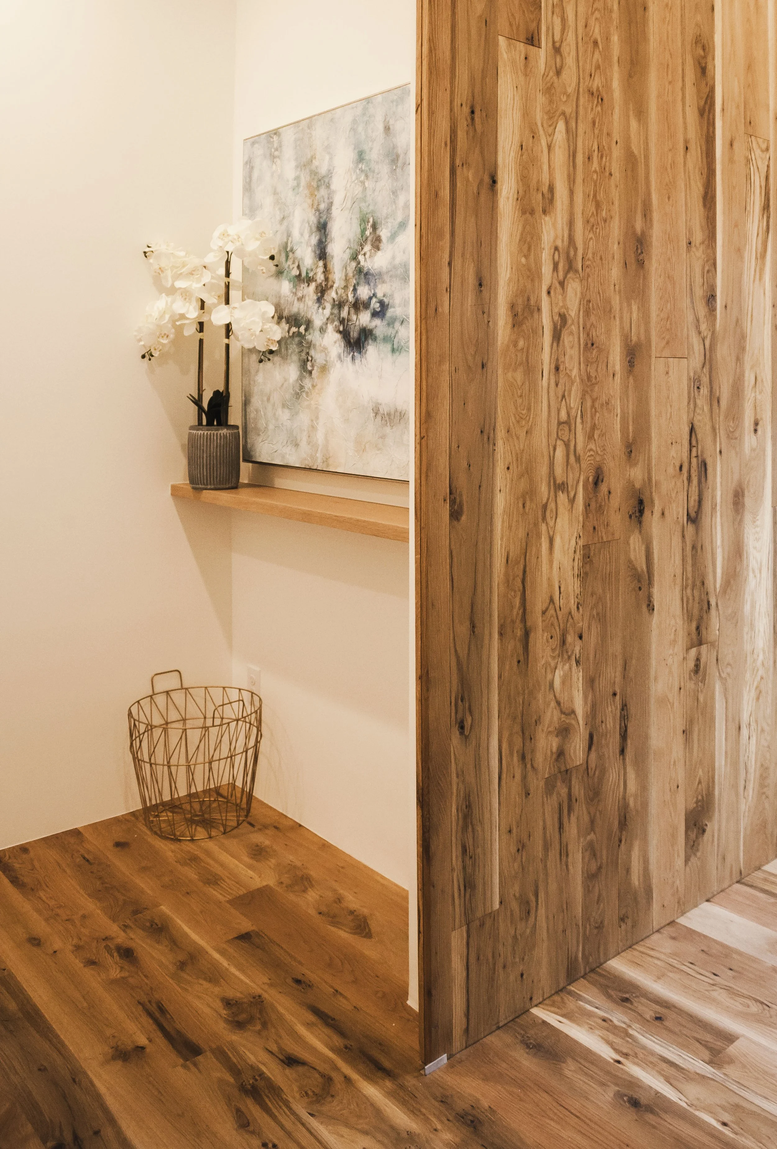 Corner of a room with wooden flooring and a partial wooden wall. A small wooden shelf holds a potted white orchid flower and a framed abstract painting. A wire basket sits on the floor.