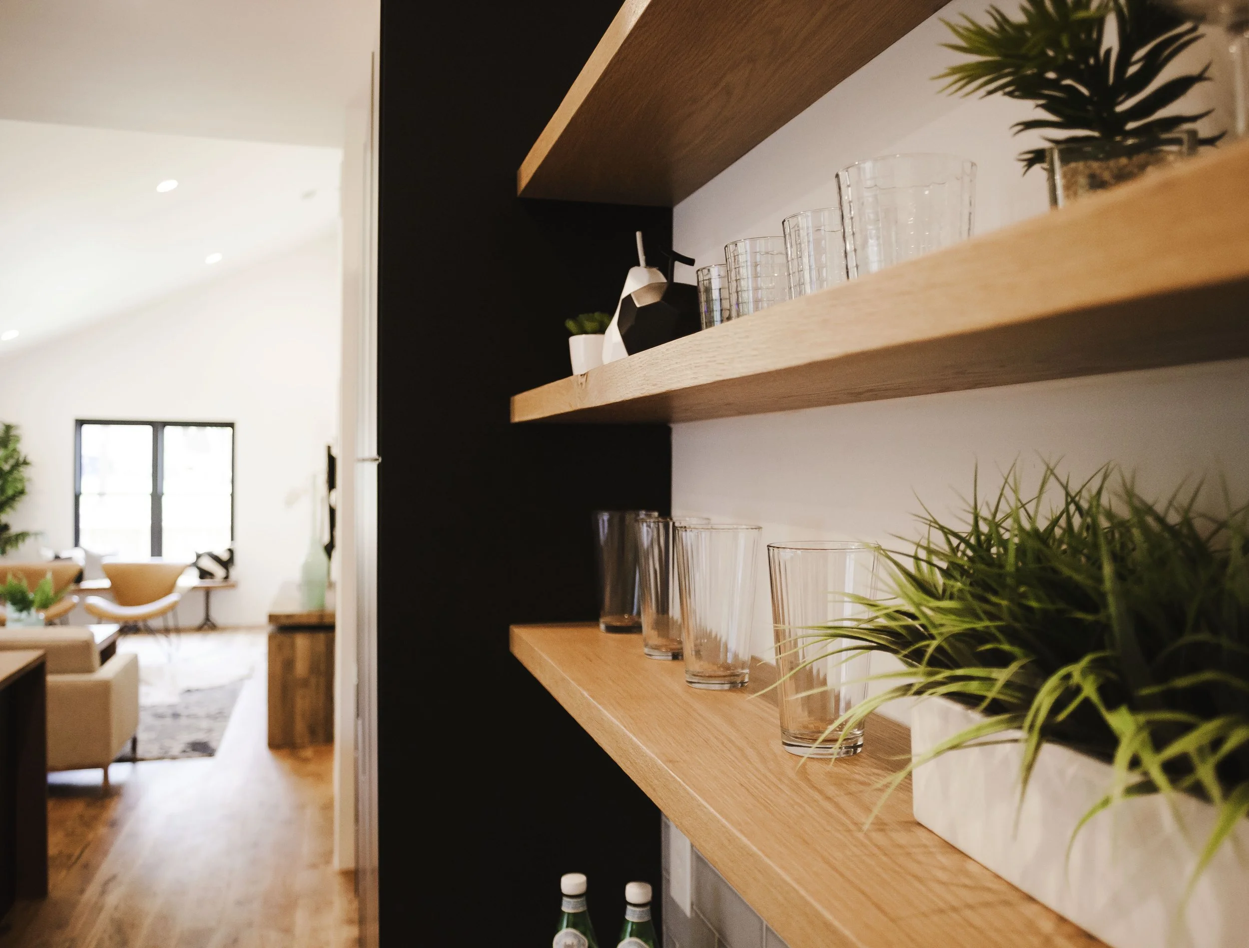 Interior view of a modern living space with wooden shelves holding glasses, potted plants, and decorative items, with a bright living room area in the background featuring chairs, large windows, and wooden flooring.