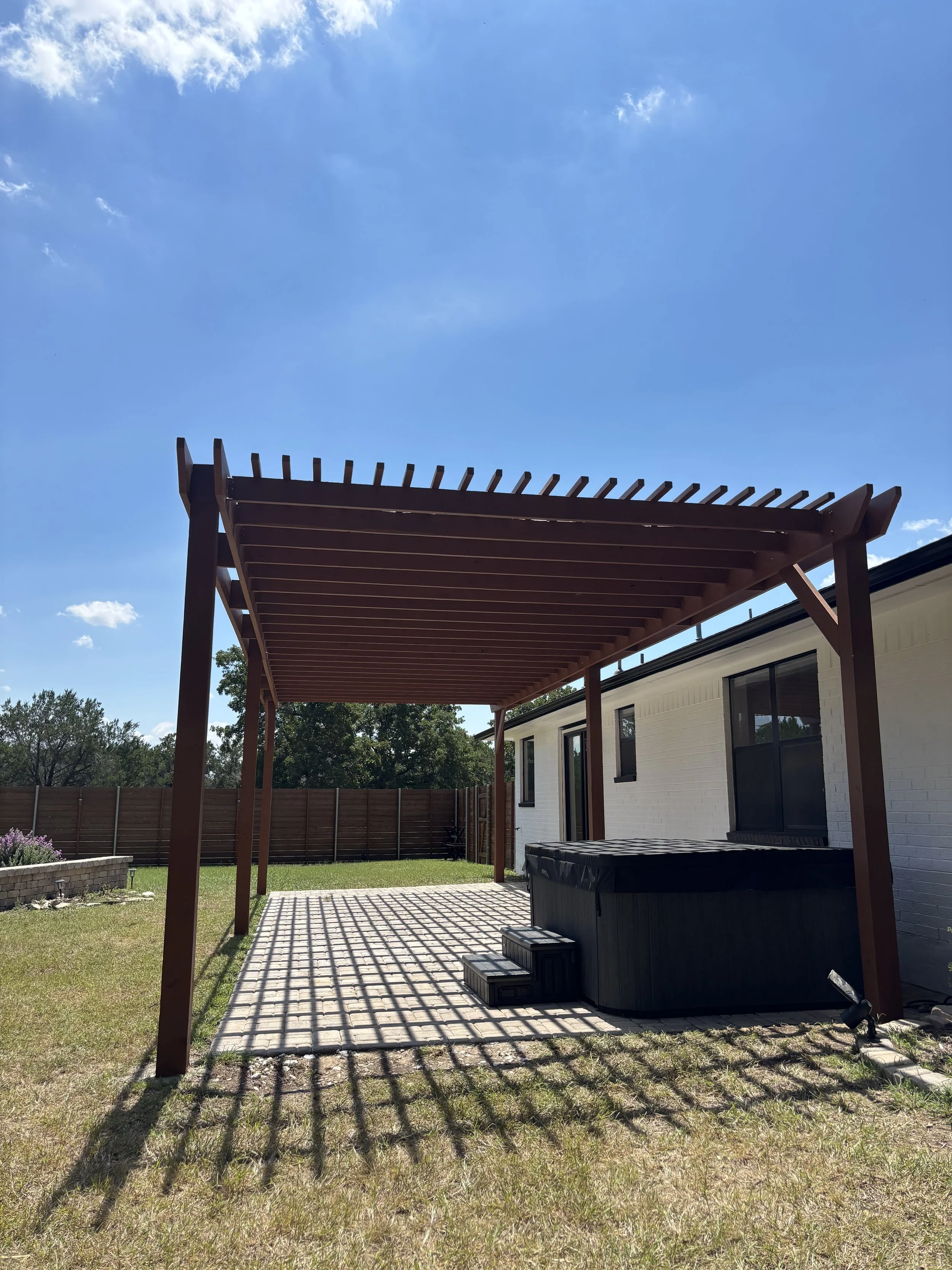 Backyard patio with a wooden pergola, hot tub, and steps, with a white house exterior and a fence in the background under a blue sky.