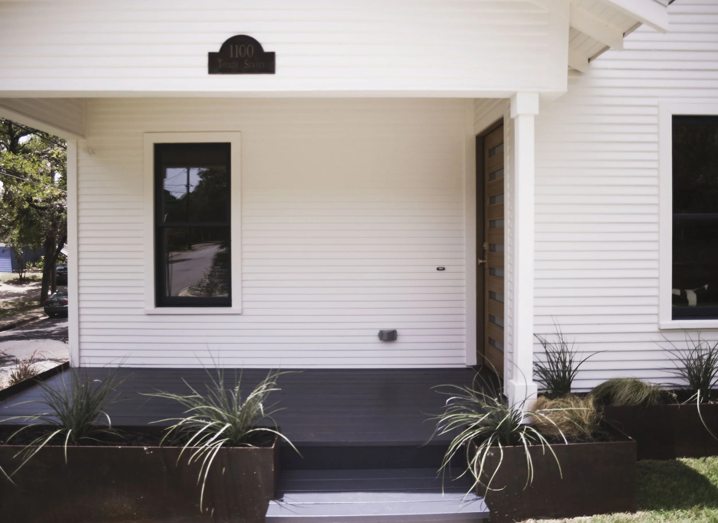 Front porch of a white house with a wooden door and a black window, decorated with black planters filled with ornamental grasses.