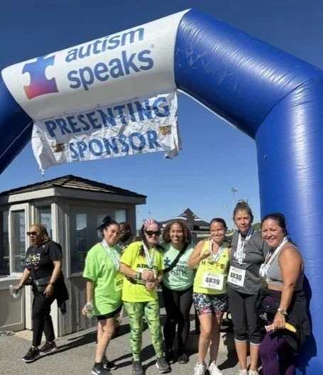 A group of 6 women runners who have medals and race numbers posed under an arch for Autism Speaks's race