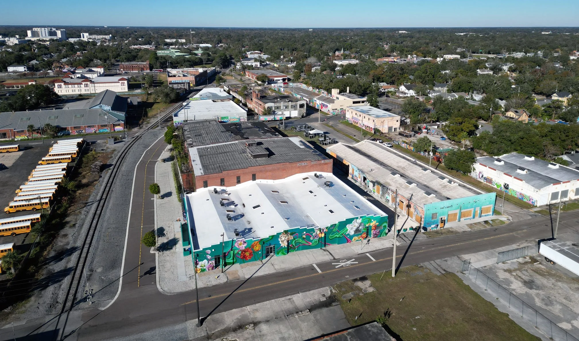 An aerial view of a row of commercial buildings with vibrant murals, a street with parked cars, and surrounding greenery, with residential houses and trees in the background.