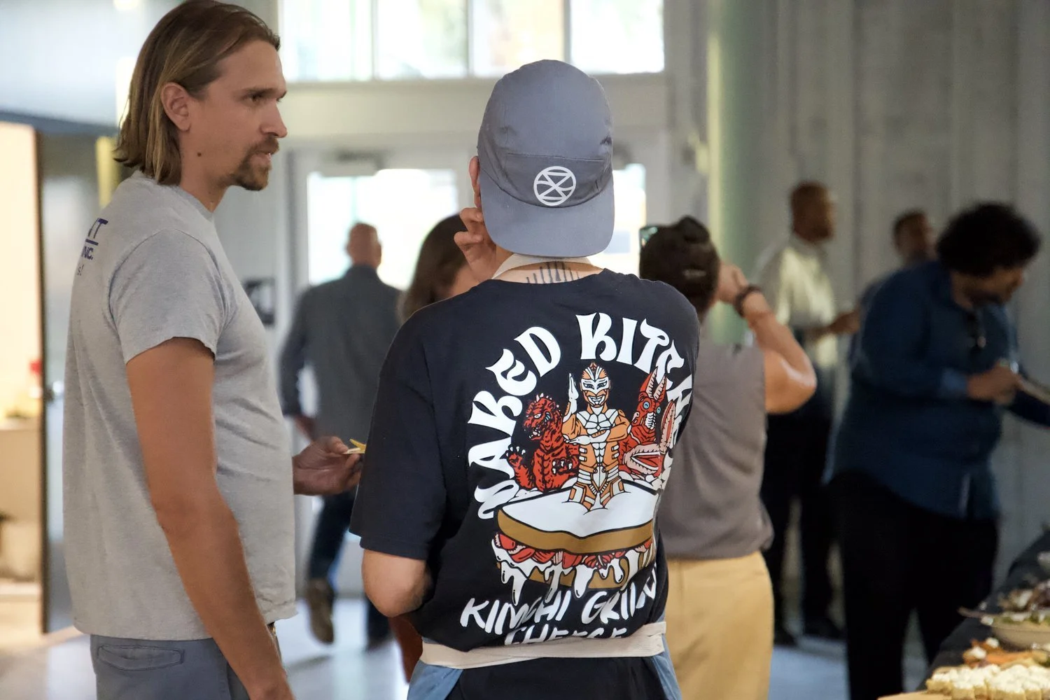 Two men conversing at a social gathering, with people in the background in an indoor space.