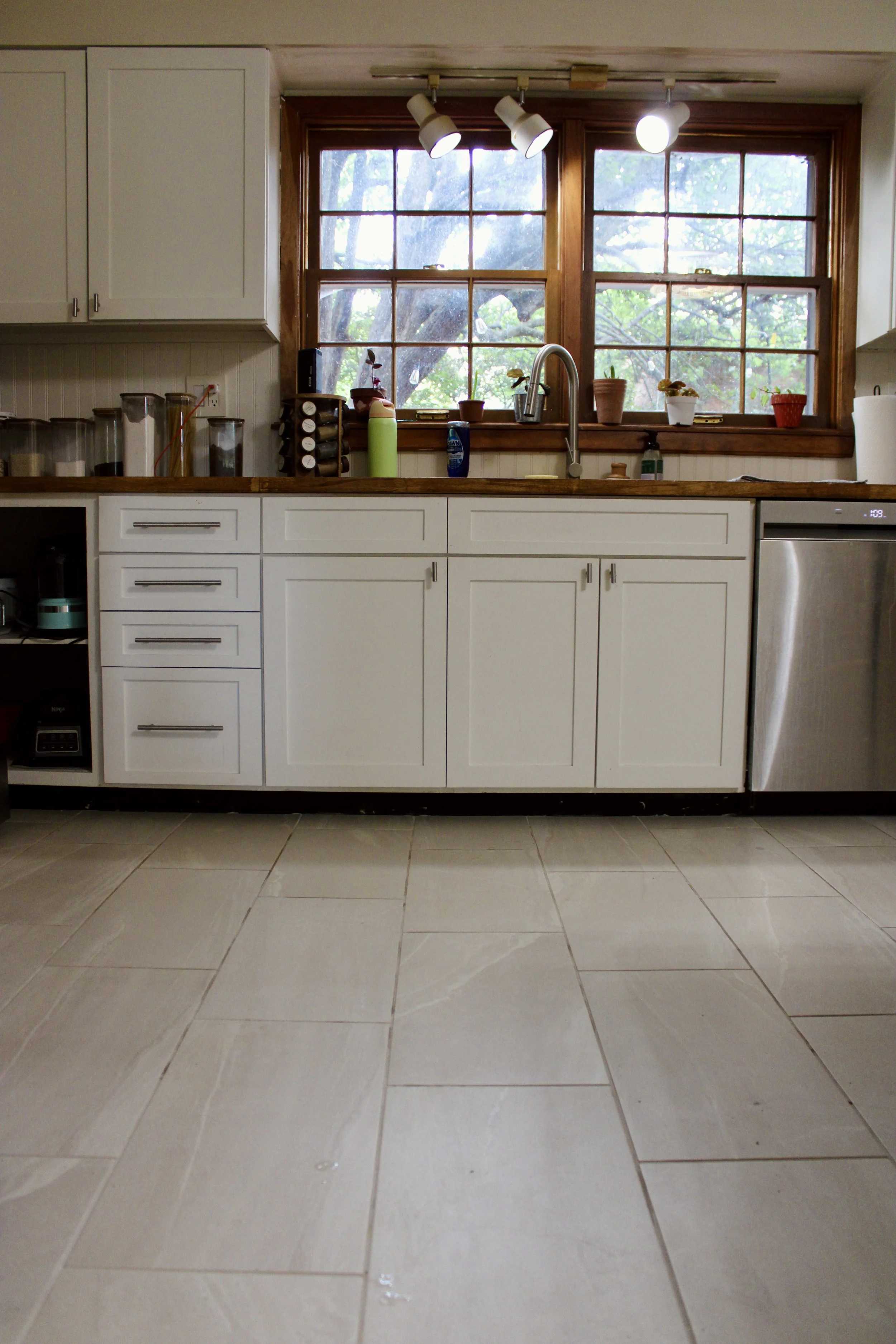 Kitchen with white-tiled backslash, open wooden shelves containing dishes, bowls, and decorative items, a window with a view of trees, a vase of purple flowers, a favorite plant, a cutting board, a coffee machine, and an oven.