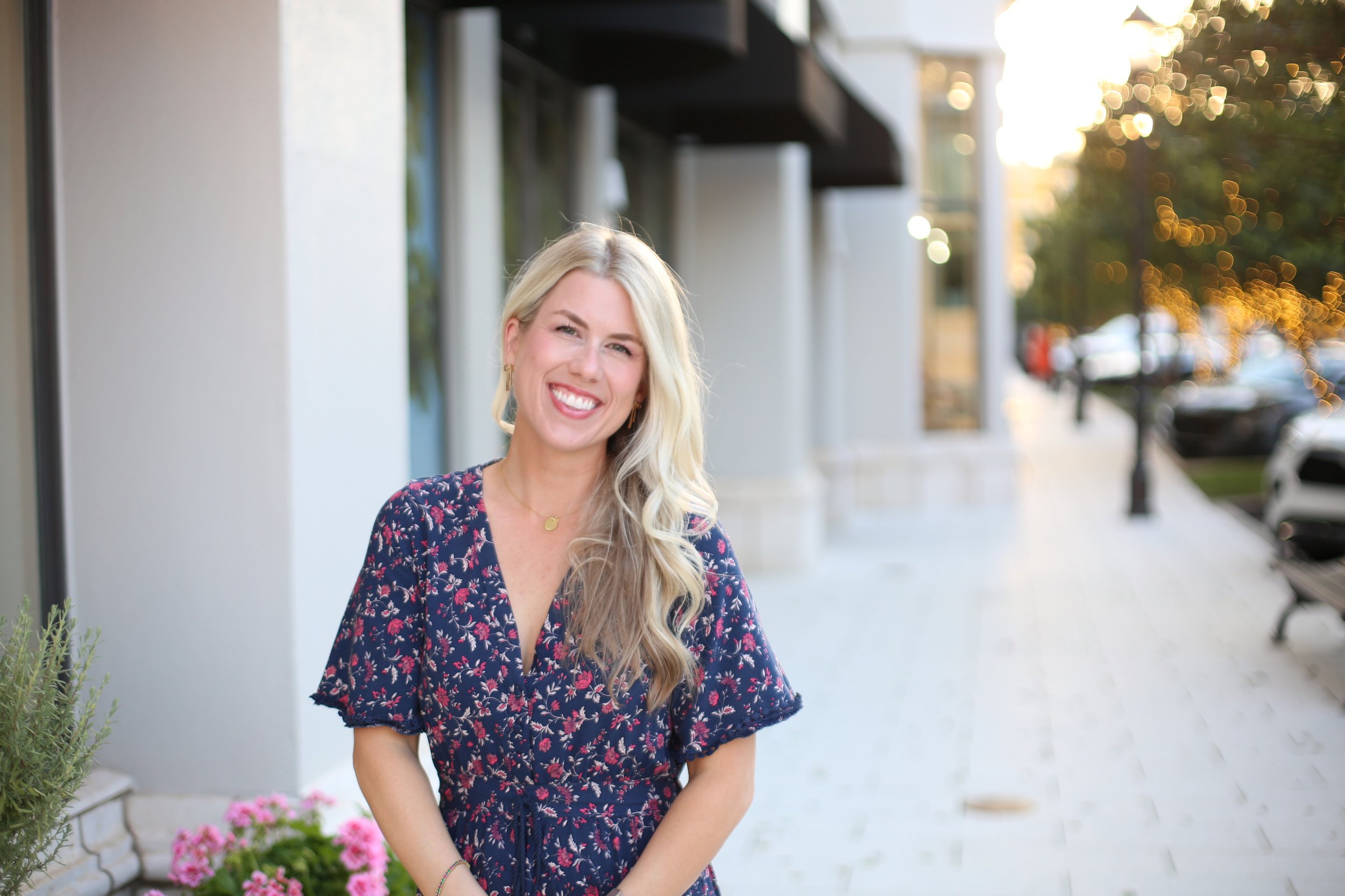 A woman with long blonde hair smiling outdoors on a city sidewalk during sunset, wearing a dark floral dress.