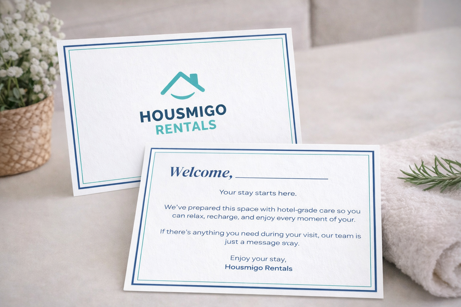 A welcome card and a rental information card from Housmigo Rentals placed on a soft white surface with a potted plant and a white towel nearby.