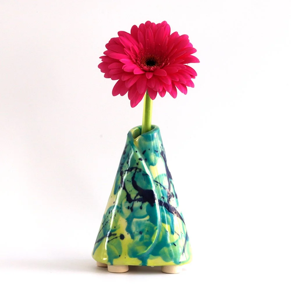A pink flower in a colorful abstract ceramic vase with black, blue, green, and yellow splashes, set against a white background.