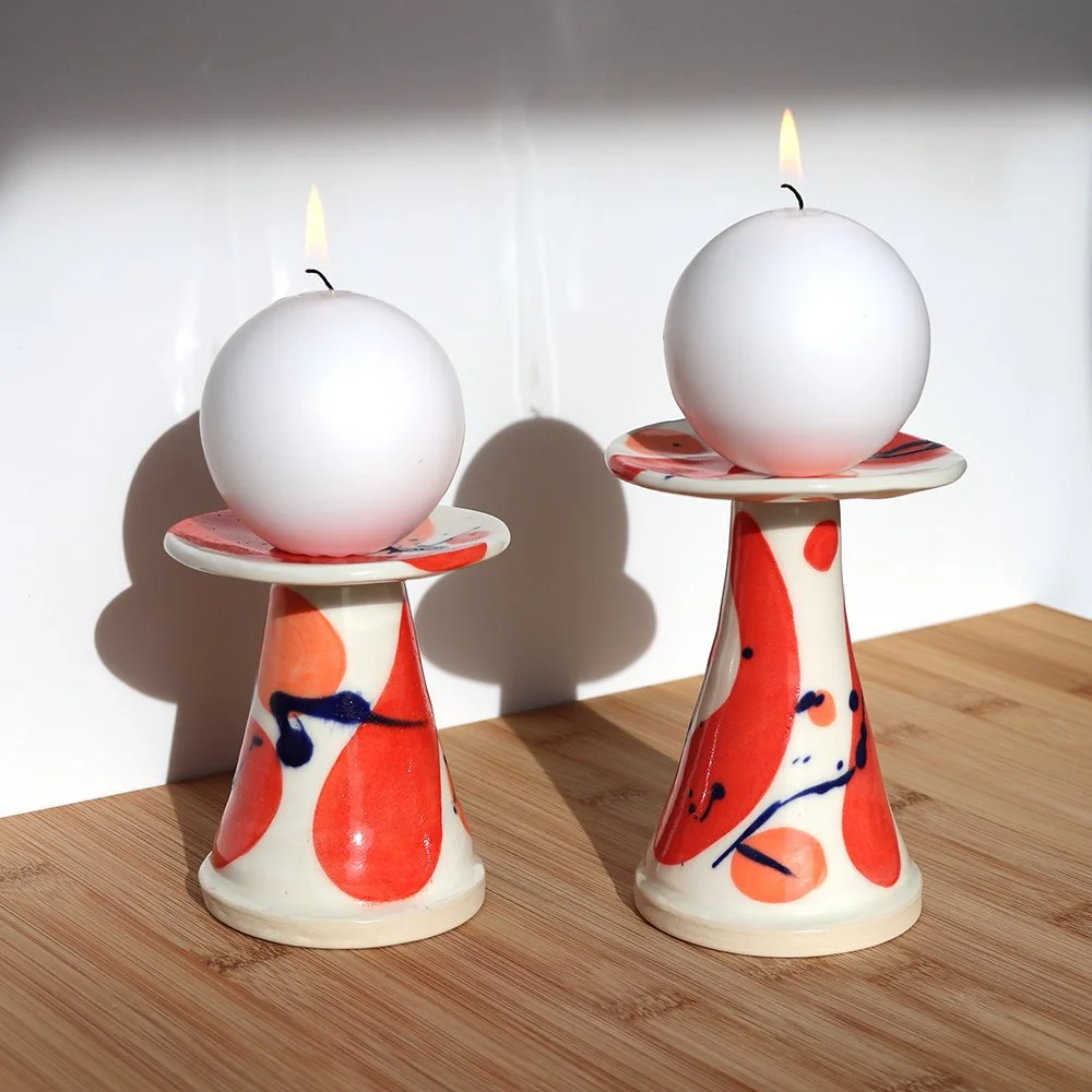 Two ceramic candle holders with abstract orange, red, and blue patterns, each holding a white spherical candle with a small lit flame, placed on a wooden surface.