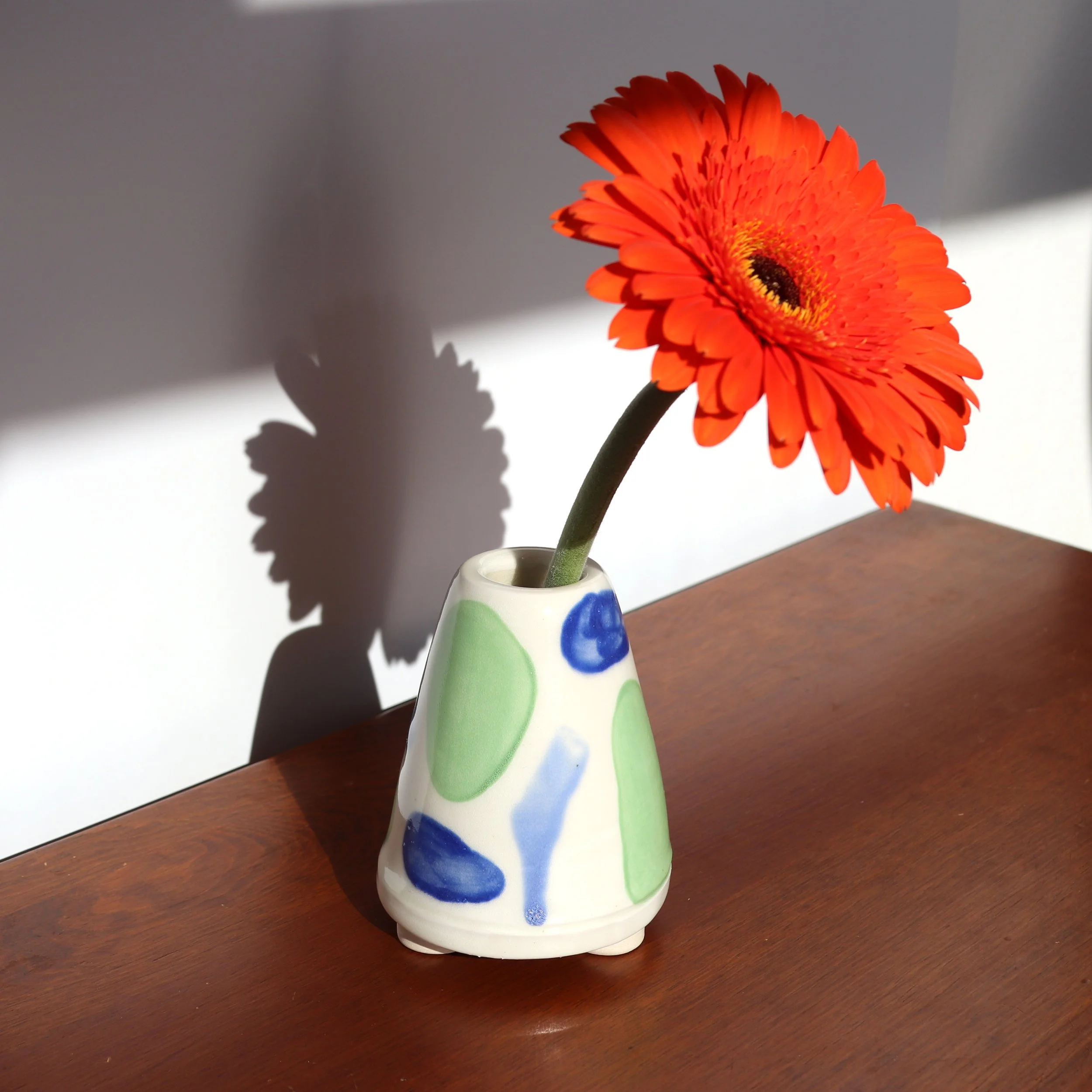 Orange gerbera daisy in a white ceramic vase with green, blue, and light blue abstract patterns, casting a shadow on a white wall, on a wooden surface.