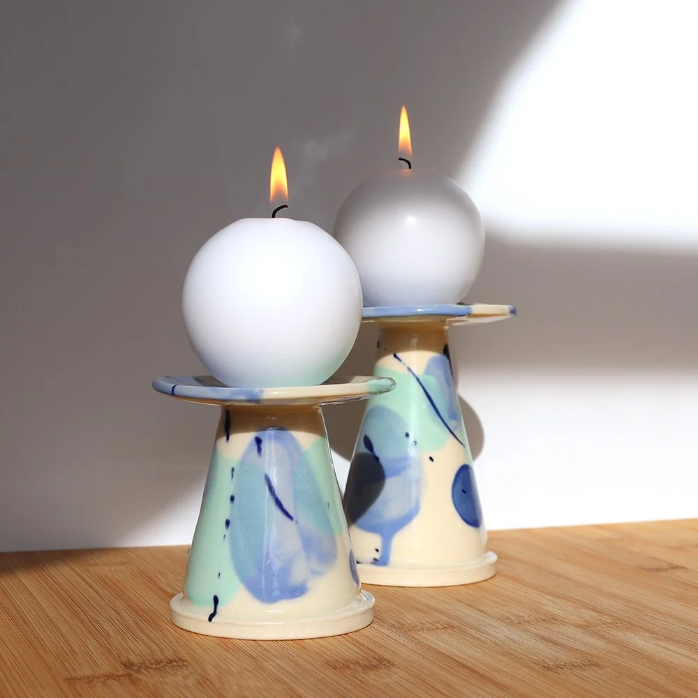 Two decorated ceramic candle holders with round candles that are lit, placed on a wooden surface near a wall with sunlight creating a shadow.