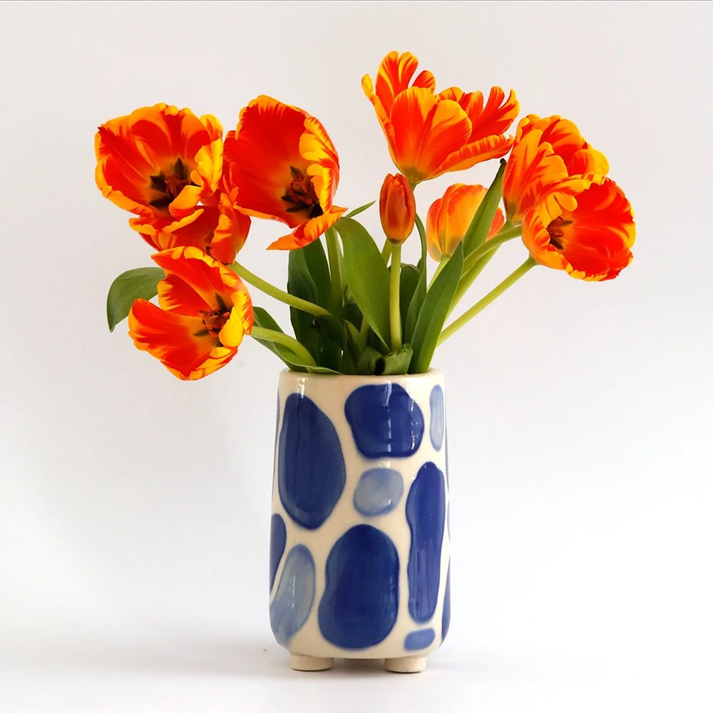 A white ceramic vase with blue abstract spots containing a bouquet of orange and yellow tulips with green stems and leaves, placed against a plain white background.