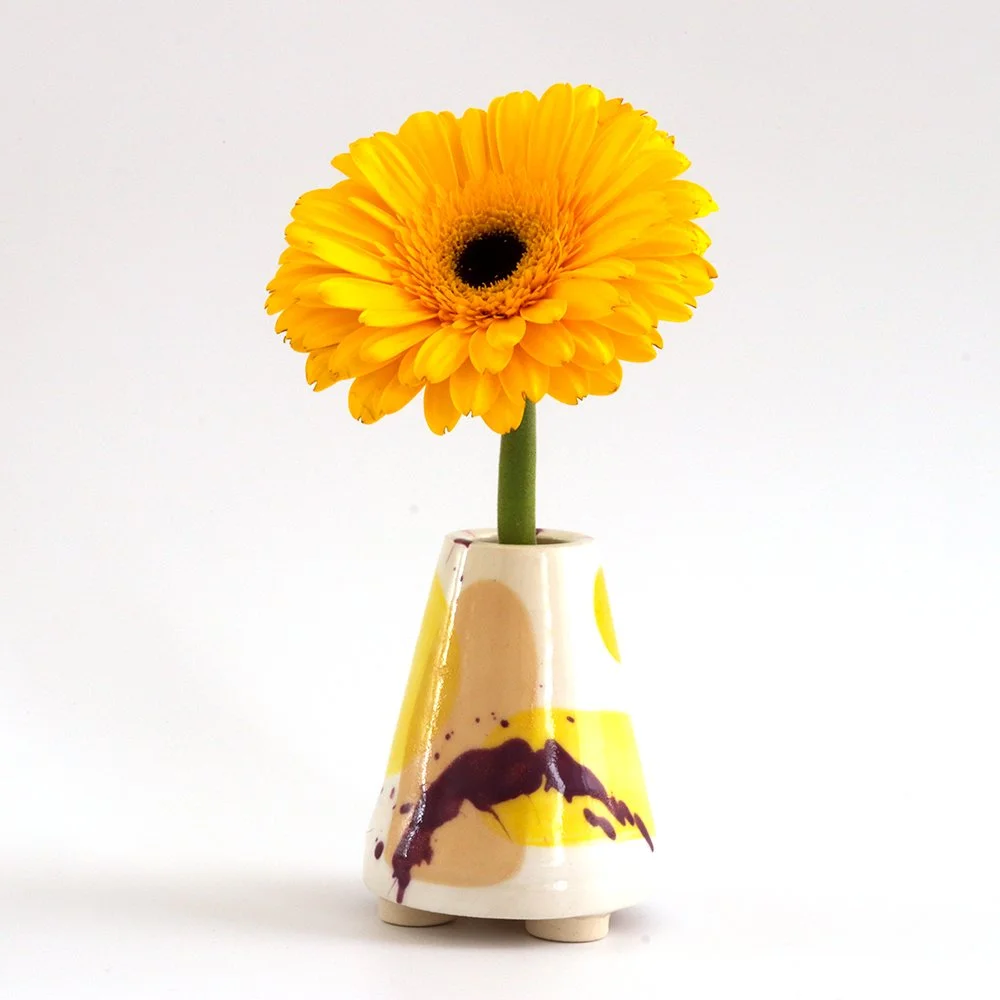 Yellow gerbera daisy flower in a ceramic vase with abstract yellow, black, and beige design, set against a white background.