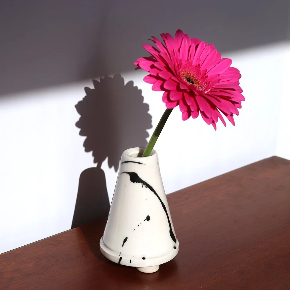 Pink gerbera daisy in a white and black ceramic vase on a wooden surface, with its shadow cast on the background wall.