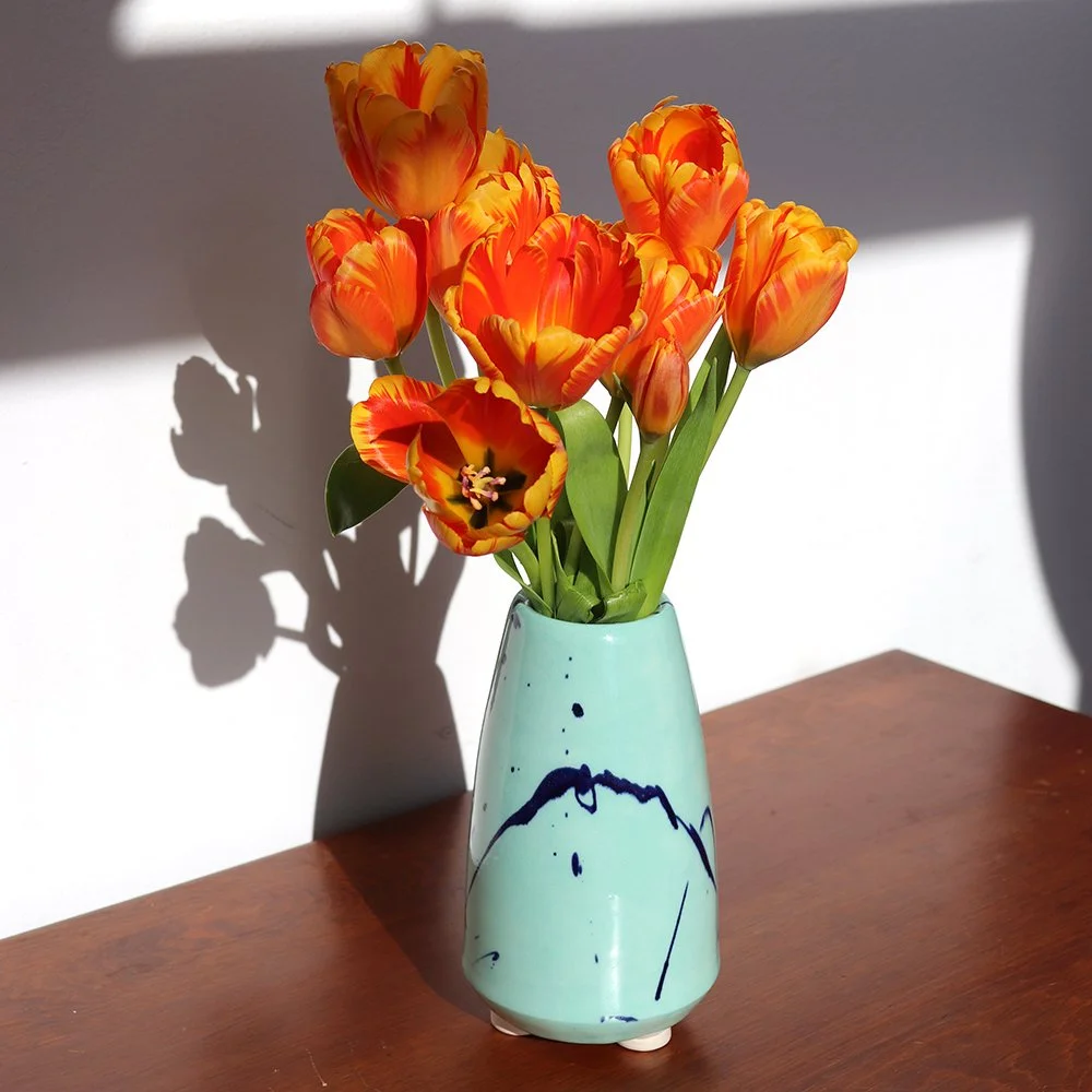 Orange and yellow tulips in a light blue vase with dark blue abstract design on a wooden surface, sunlight casting a shadow on a white wall.