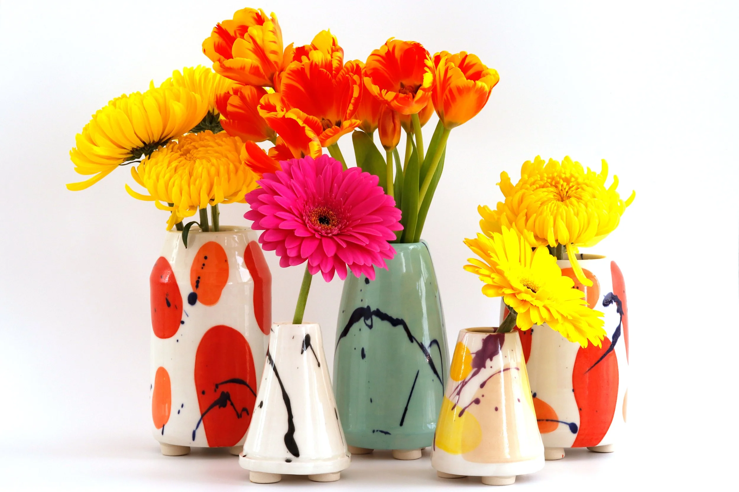 Colorful flowers in patterned vases, including pink gerbera, yellow chrysanthemums, and orange tulips, arranged on a white background.