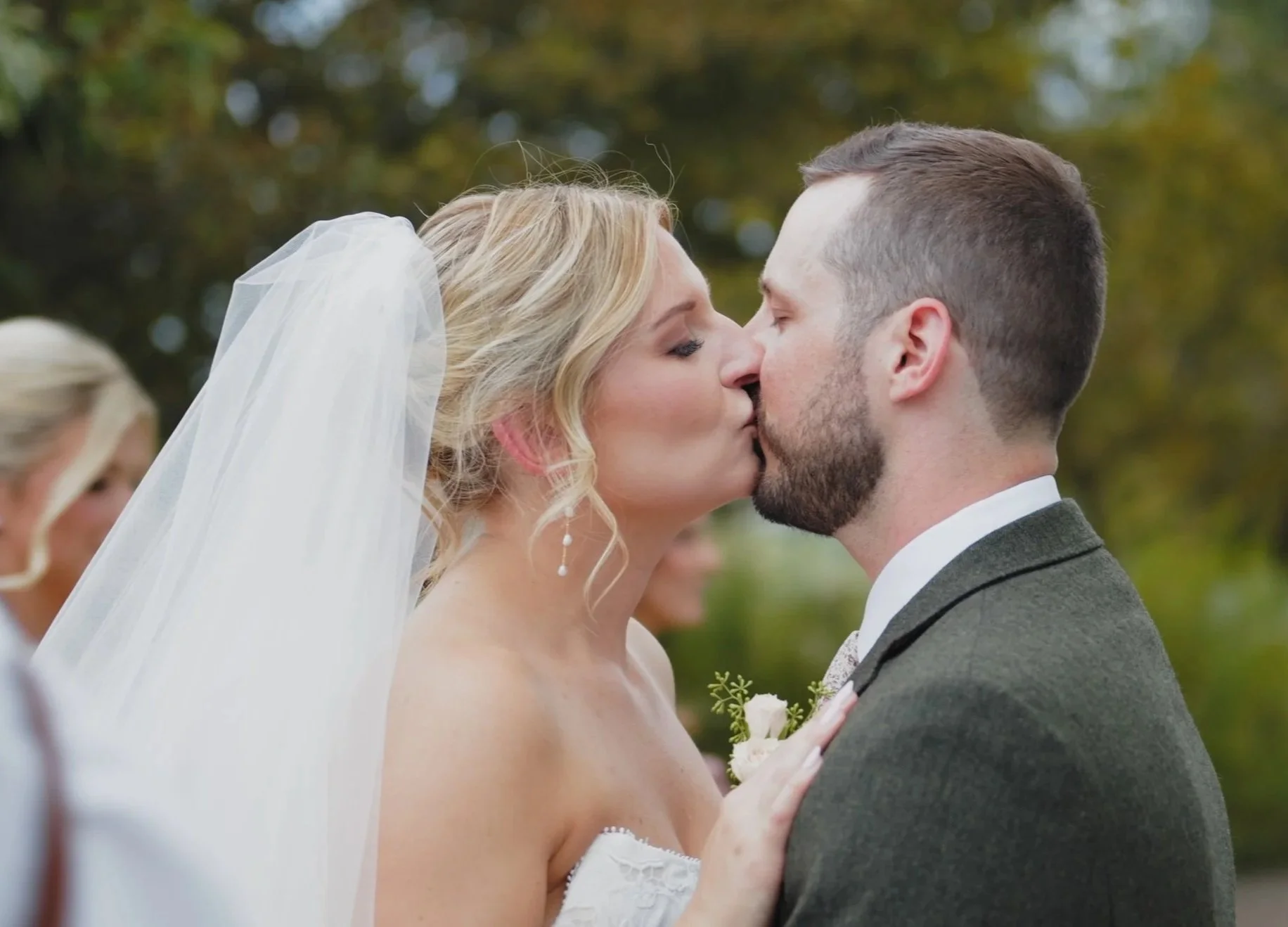A bride and groom kissing outdoors during their wedding ceremony, with the bride wearing a white veil and wedding dress, and the groom in a dark suit and tie.