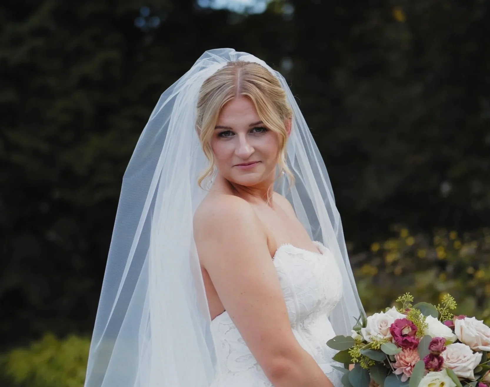A bride in a strapless white wedding dress and veil holding a bouquet of pink, white, and purple flowers outdoors.