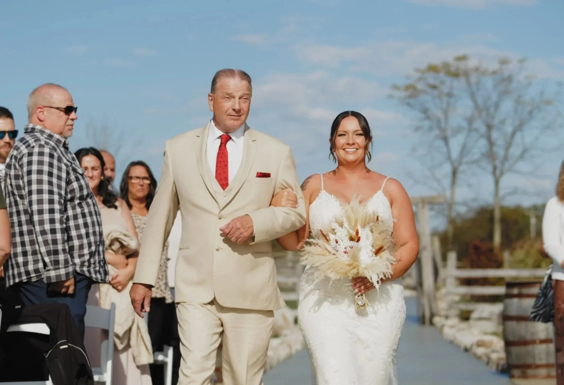 A bride in a white wedding dress holding a bouquet walking outdoors with an older man in a light suit. People are standing on either side of the walkway, watching the procession. The background shows trees and a cloudy sky.