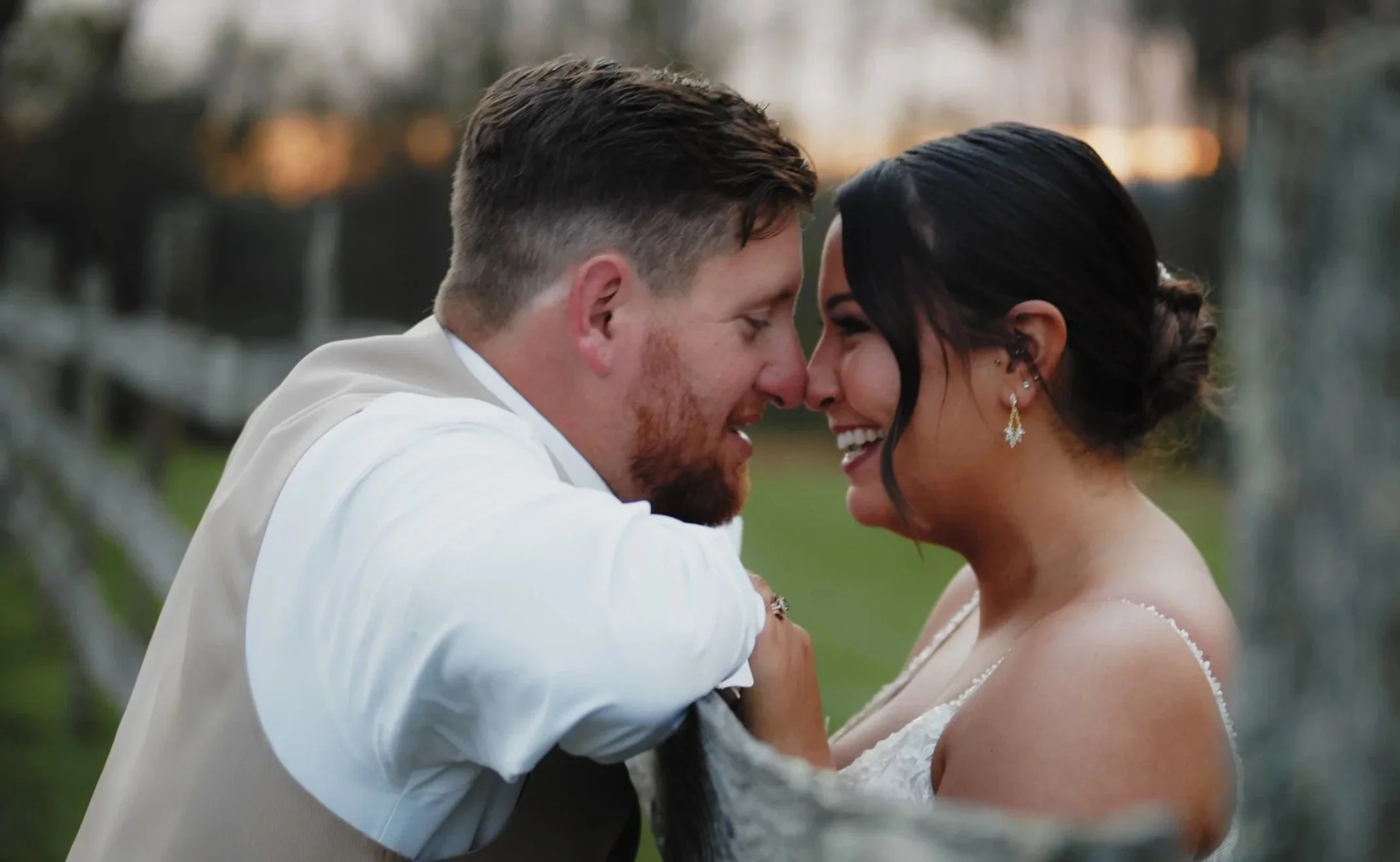 A couple smiling and touching foreheads outdoors during sunset, with a blurred background of trees and sky.