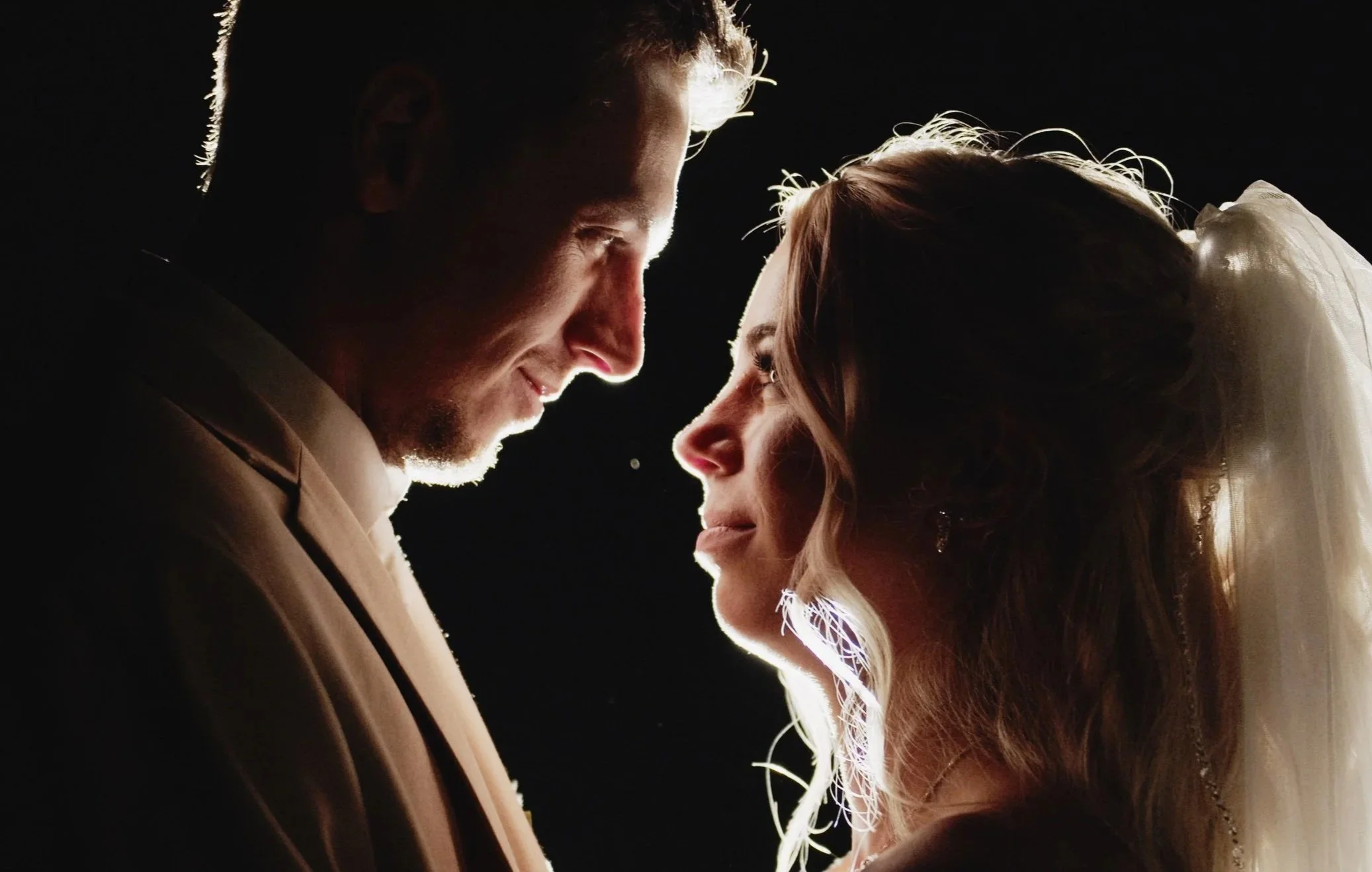 A couple in wedding attire, facing each other closely against a black background, with backlighting highlighting their profiles.