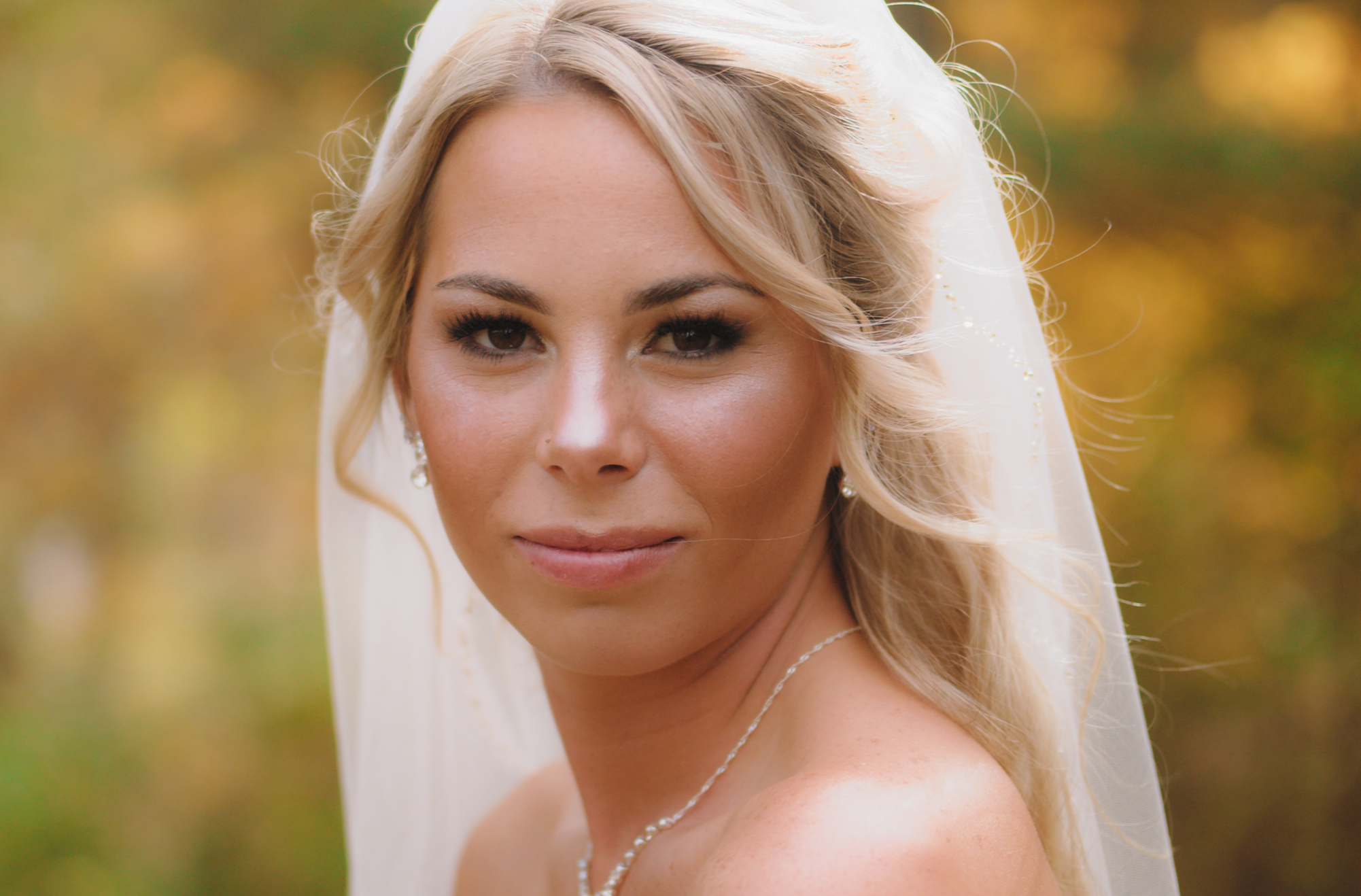 Close-up of a blonde woman in wedding attire with a white veil, outdoor background with trees and autumn foliage.