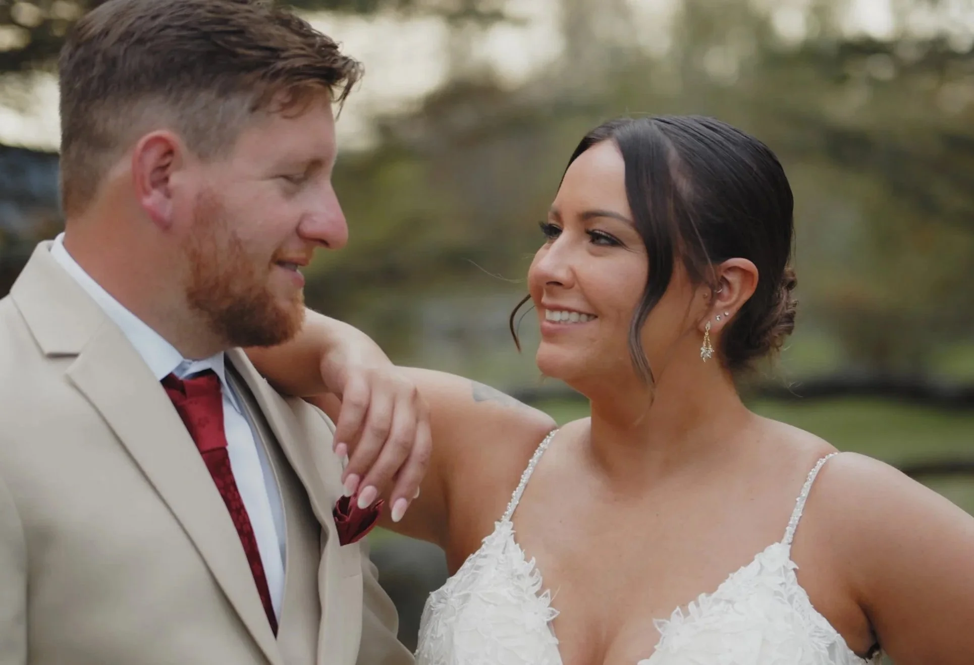 A newlywed couple looks at each other lovingly outdoors during sunset, with the groom in a beige suit and the bride in a white wedding dress with lace details.