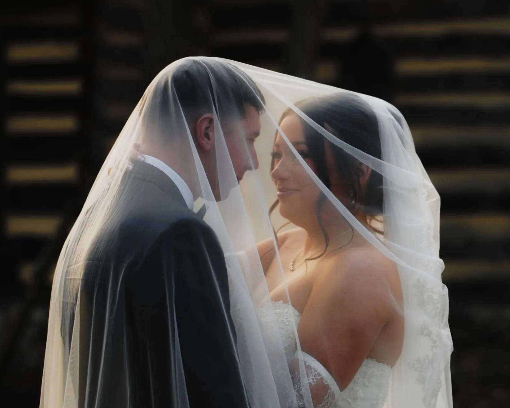 A bride and groom standing close together with a sheer veil overlapping their faces, smiling at each other on their wedding day.