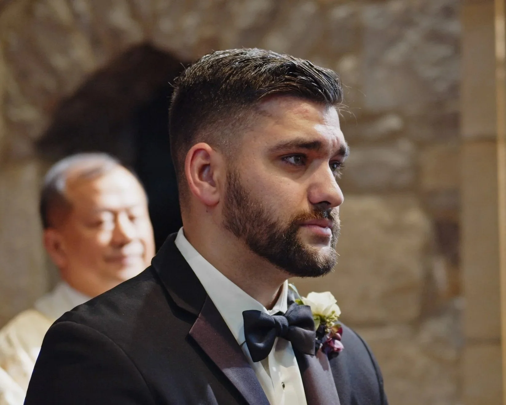 A groom with dark hair and beard, wearing a black tuxedo, white shirt, and black bow tie, standing during a wedding ceremony, with a man in the background smiling.
