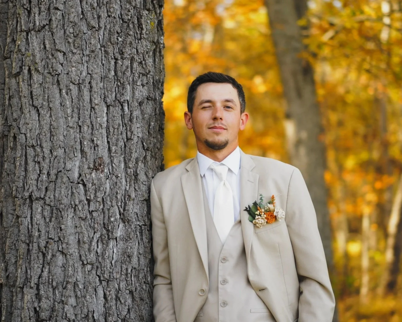 A man in a beige suit with a boutonniere, standing in front of a large tree in an autumn forest, winking at the camera.