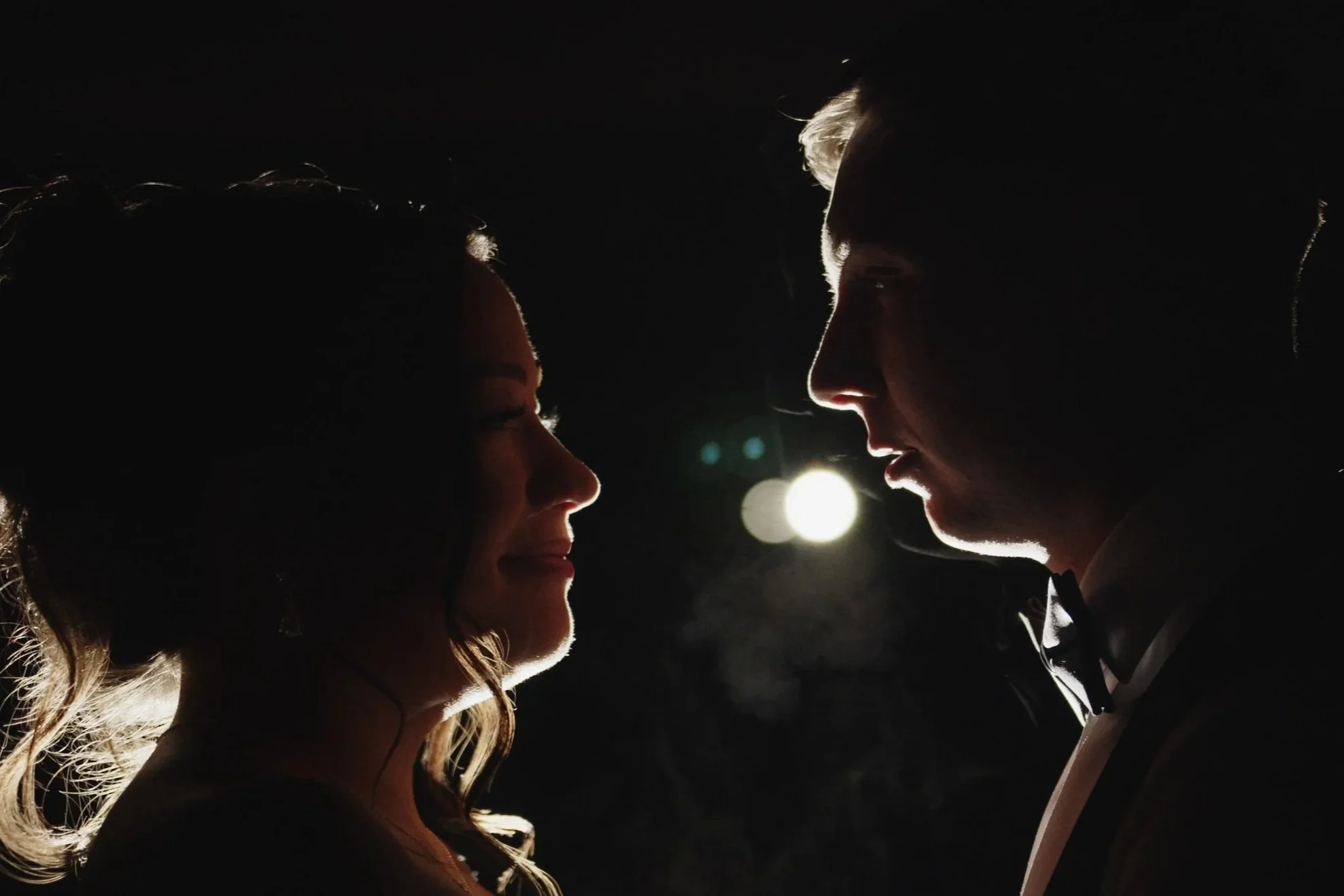 Silhouettes of a wedding couple facing each other in a dark setting with backlighting, highlighting their profiles.