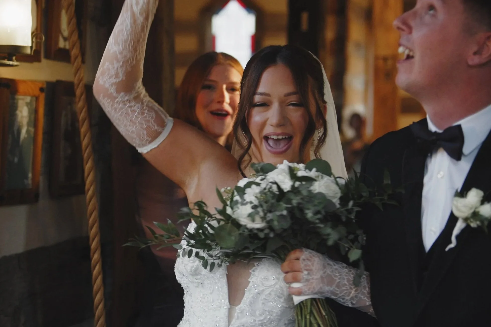 Joyful bride holding a bouquet of white flowers and greenery, surrounded by happy attendees, at her wedding celebration.
