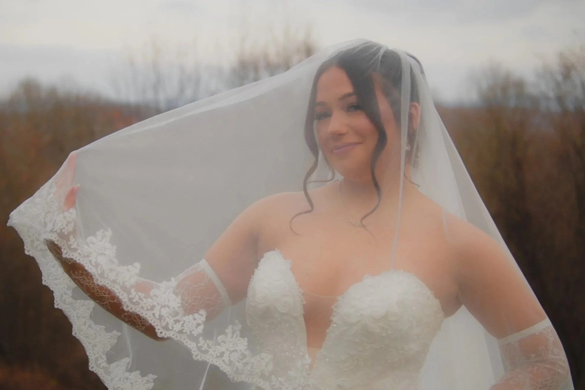 A bride wearing a white wedding dress and veil outdoors, smiling at the camera, with blurred trees in the background.