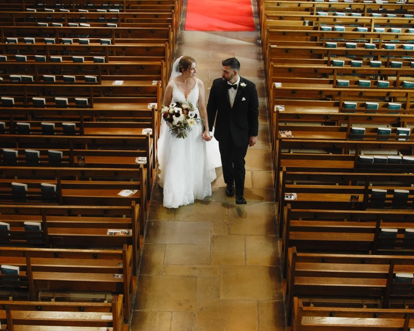 A bride and groom walking hand in hand down the aisle of a church. The bride is wearing a white wedding dress and holding a large bouquet of flowers, and the groom is dressed in a black tuxedo with a bow tie. The church has wooden pews and a red carp