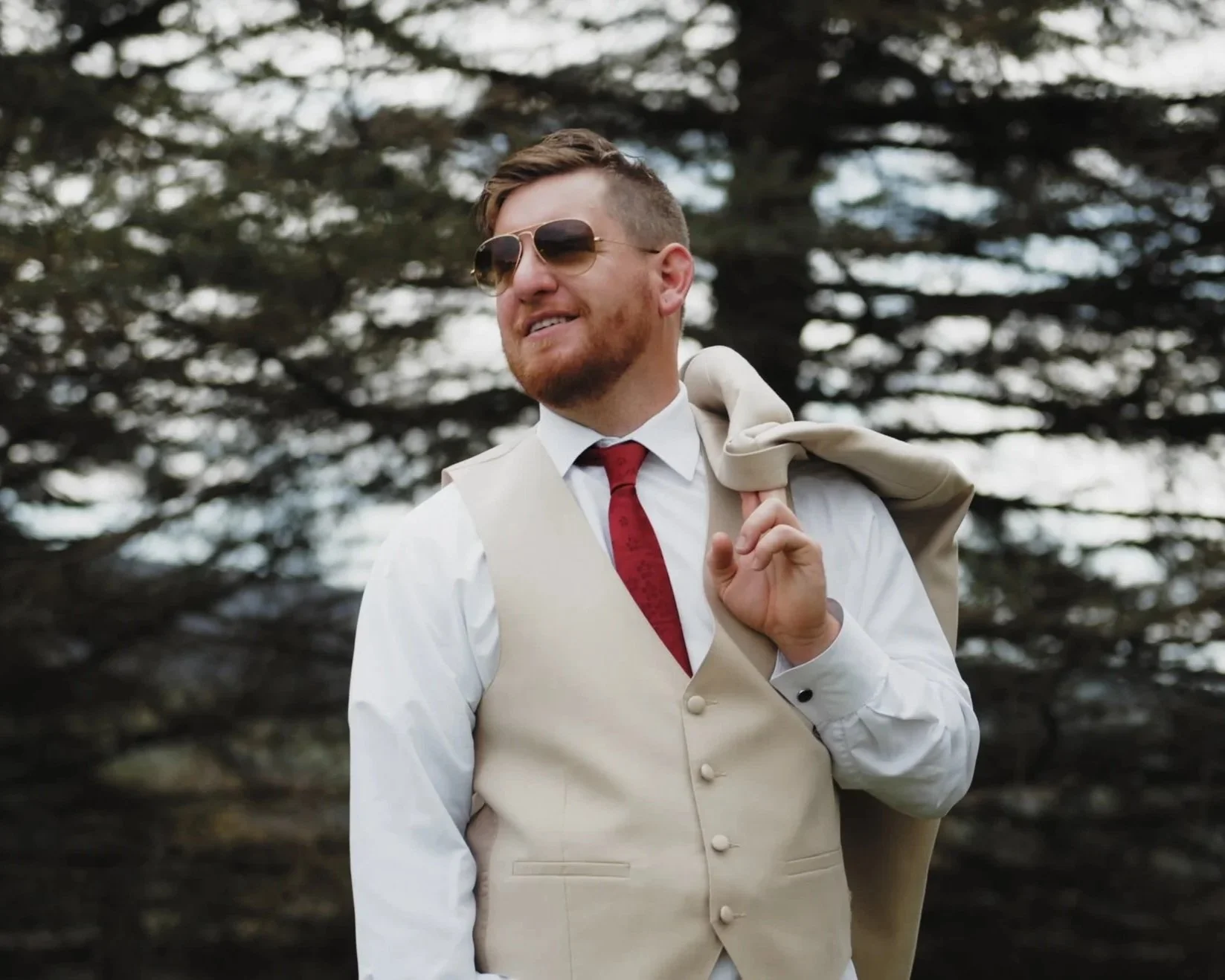 A man in a white dress shirt, beige vest, and red tie, carrying a beige jacket over his shoulder, standing outdoors in front of a large tree, wearing sunglasses, and smiling.