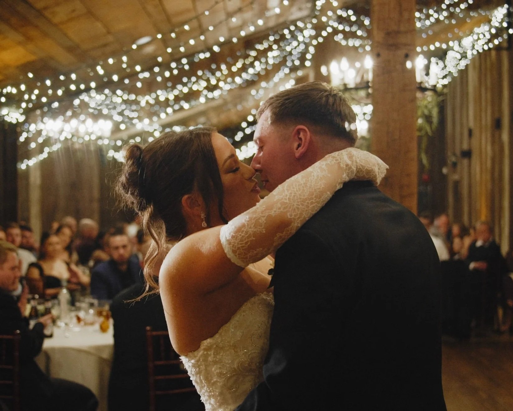 A newlywed couple sharing a dance at their wedding reception, surrounded by guests, with string lights hanging from the ceiling.
