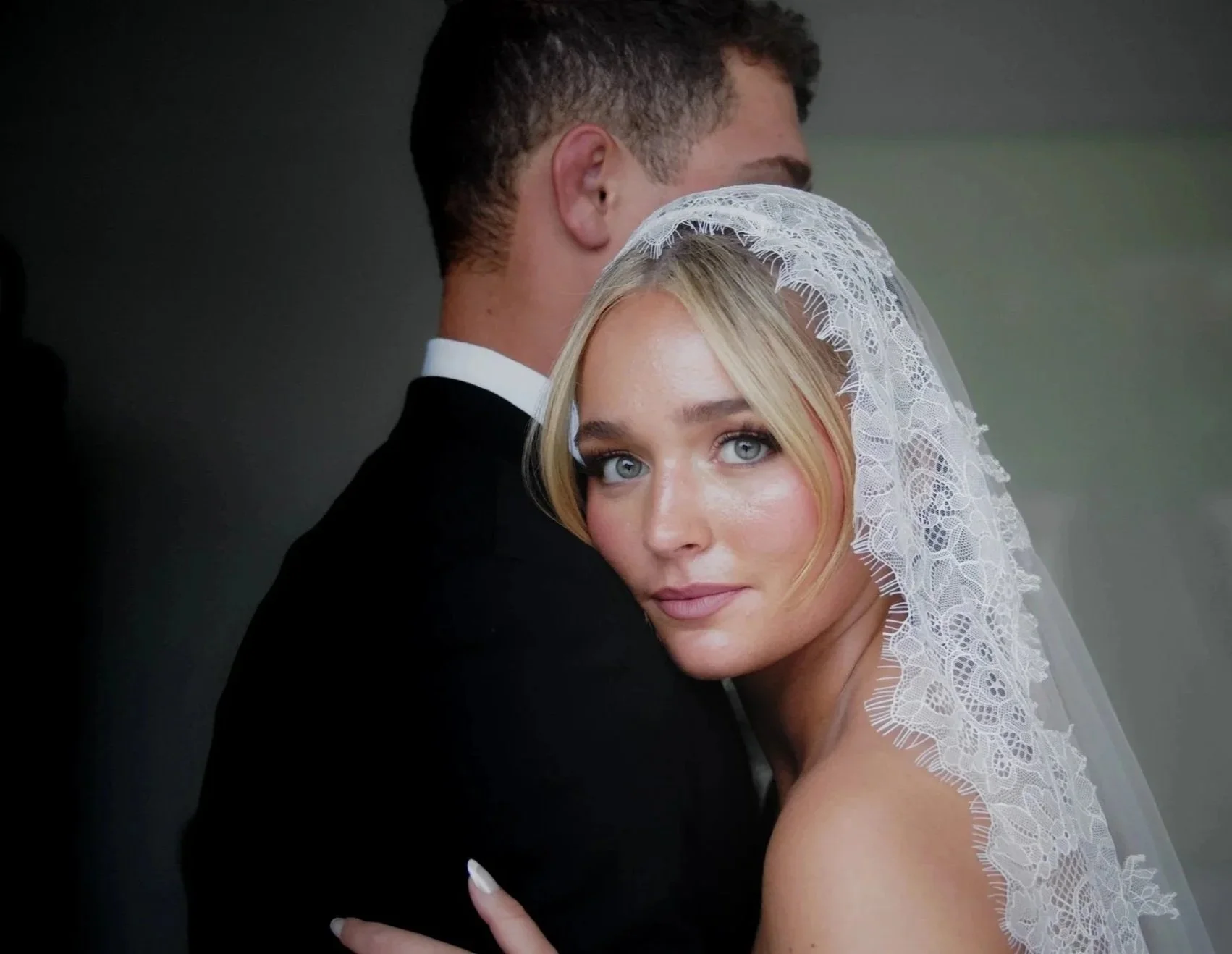 A bride in a lace veil hugging a groom in a tuxedo, with her face turned towards the camera and blue eyes visible.