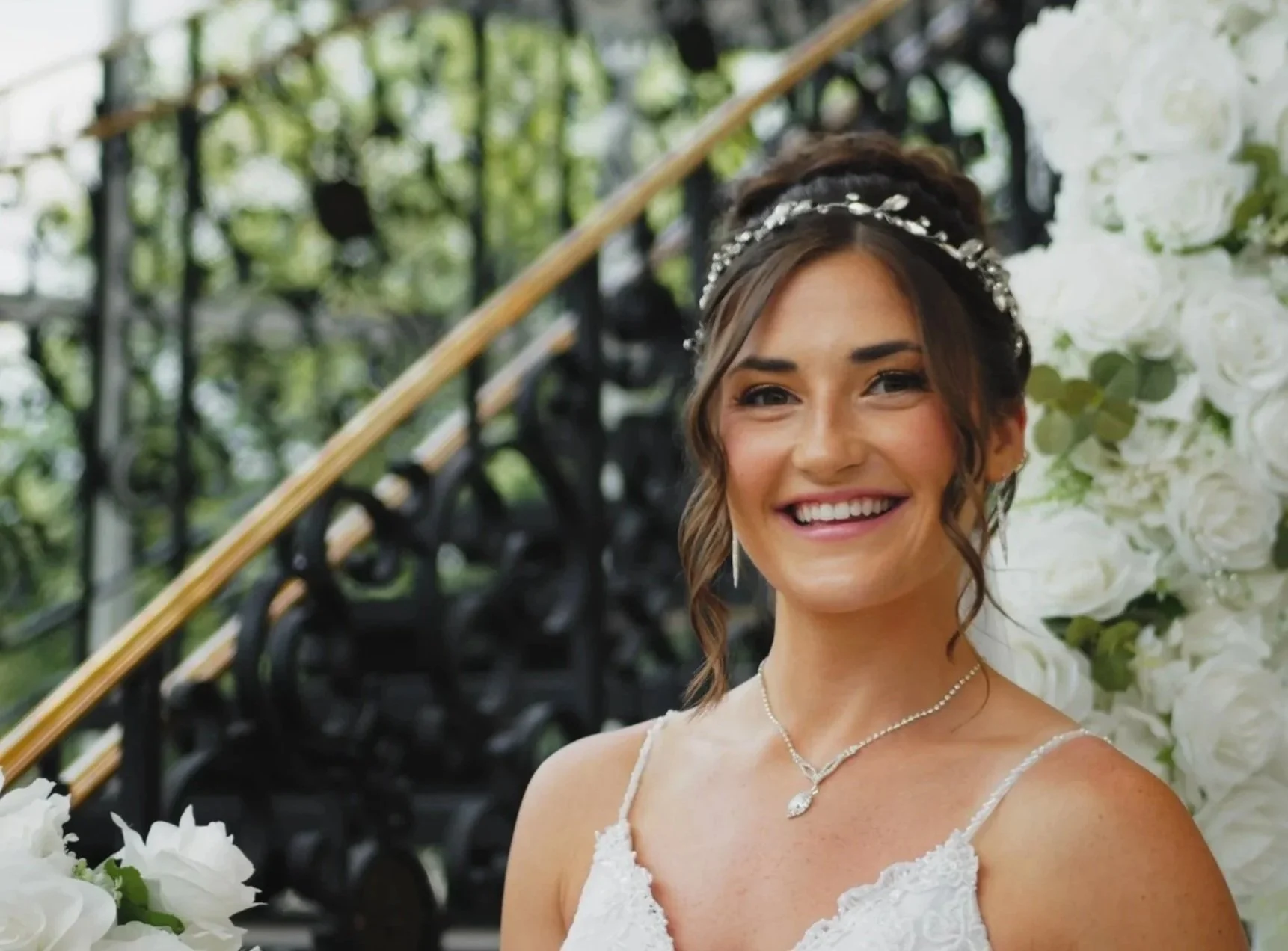 A smiling woman in a wedding dress, wearing a tiara and necklace, standing next to a floral backdrop of white roses and greenery.