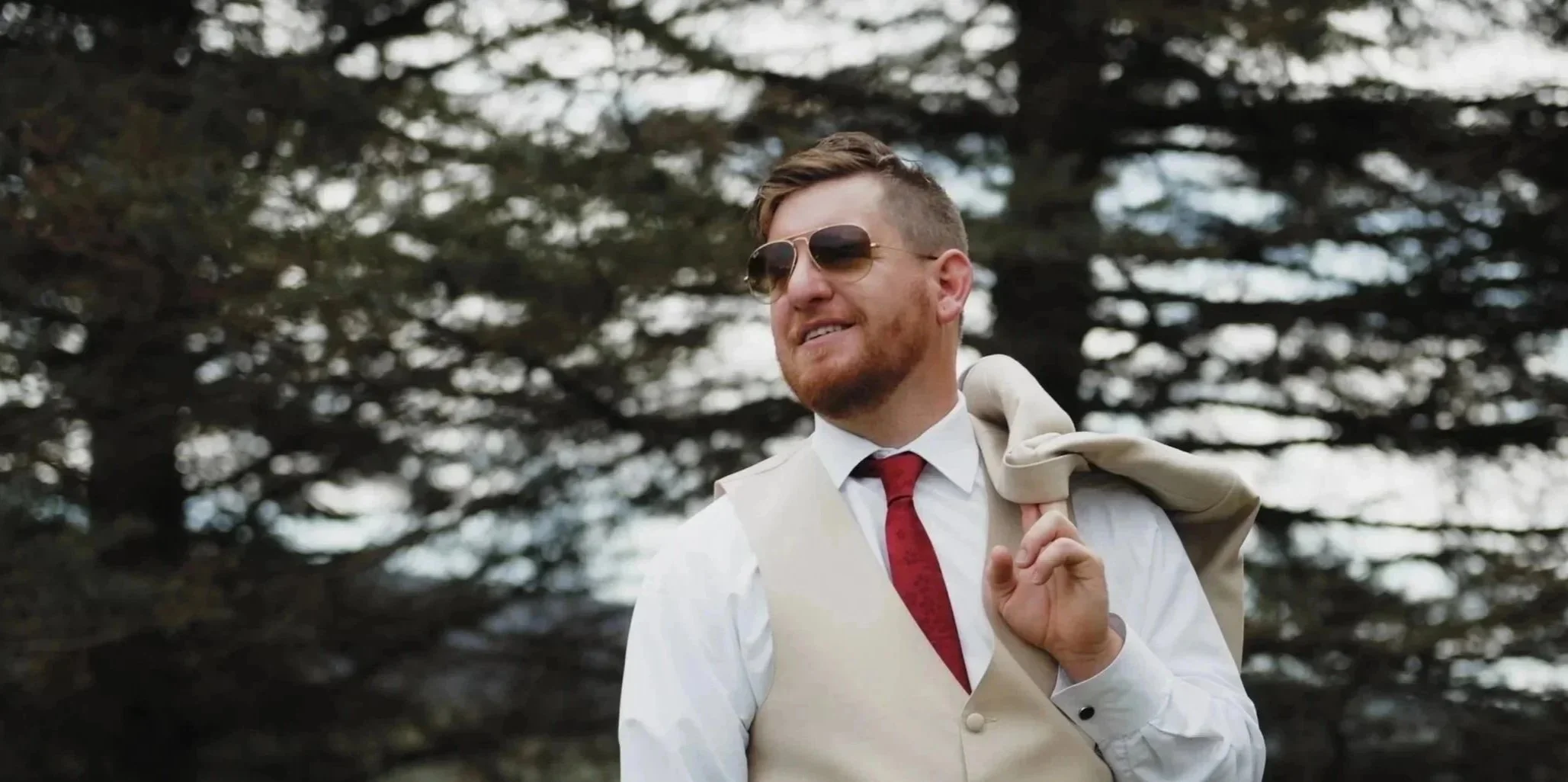 A man with brown hair and a beard, wearing sunglasses, a white shirt, a beige vest, a red tie, and holding a beige jacket over his shoulder, standing outdoors with trees in the background.