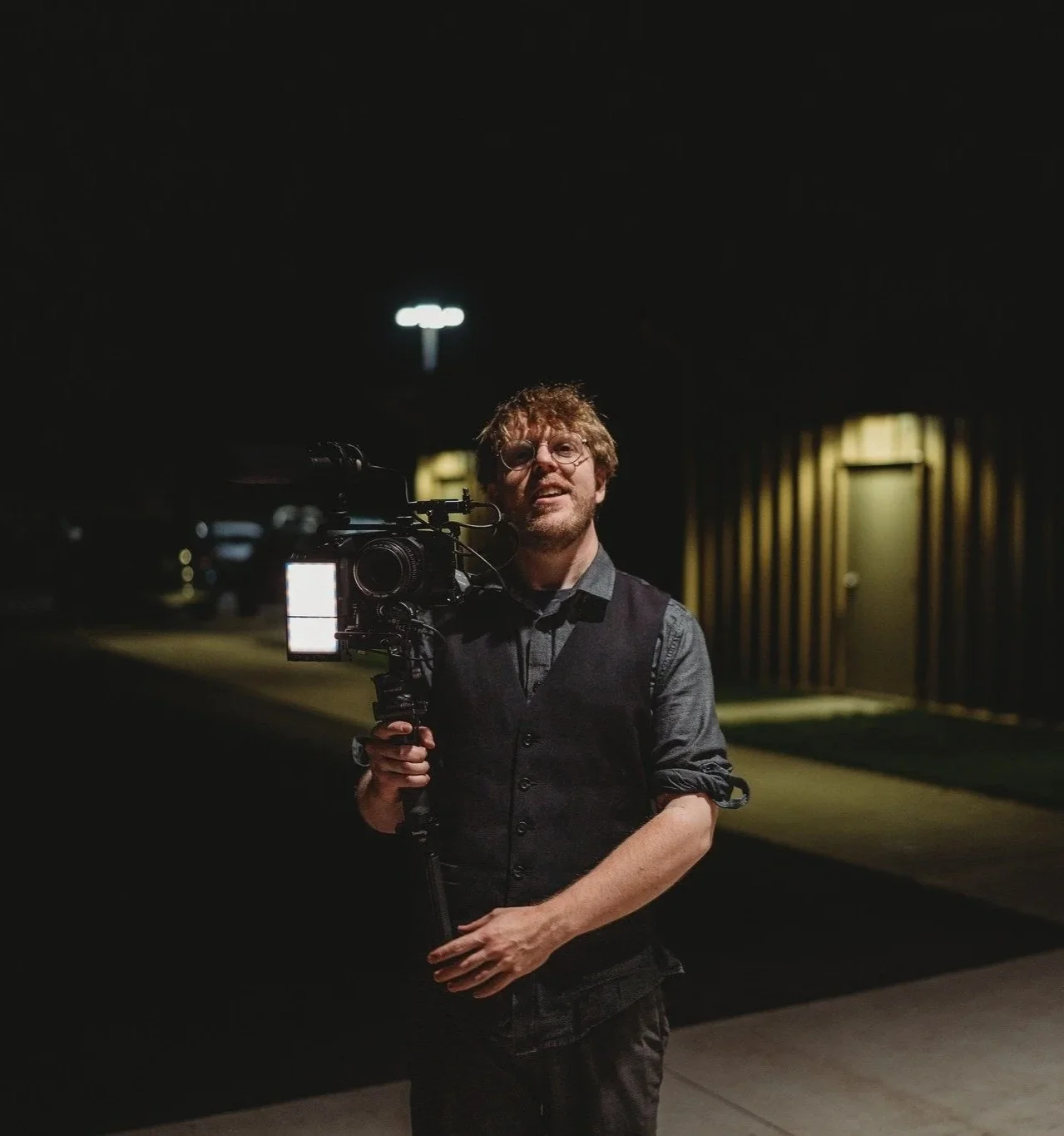 A man with curly hair, glasses, and a beard standing outdoors at night, holding a professional camera with a microphone and light attached, illuminated by a nearby streetlamp, with a small building in the background.