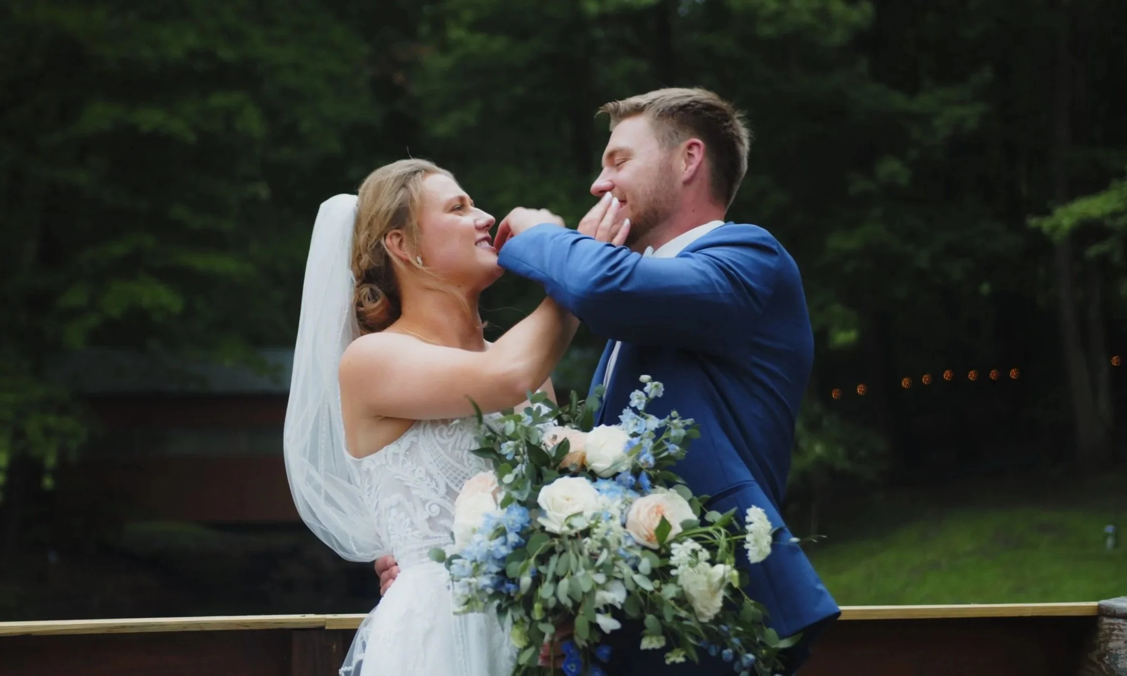 A bride and groom share a joyful moment outdoors, the bride holding a bouquet of white and blue flowers, with trees in the background.