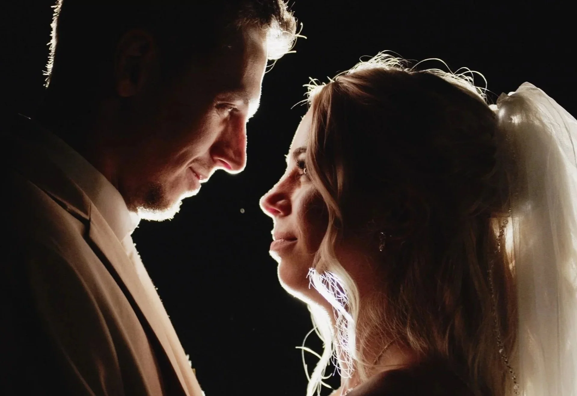 A man and woman face each other closely against a black background, with lighting highlighting their profiles. The woman is wearing a wedding veil and earrings, and the man is in a suit.